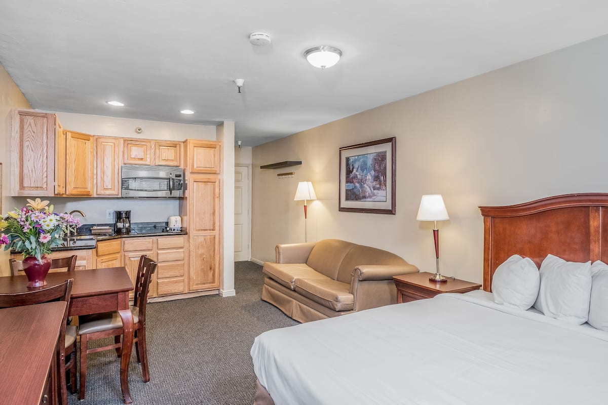 A hotel room with a bed, a sofa, a dining table with chairs, a kitchen area, and a framed picture on the wall.