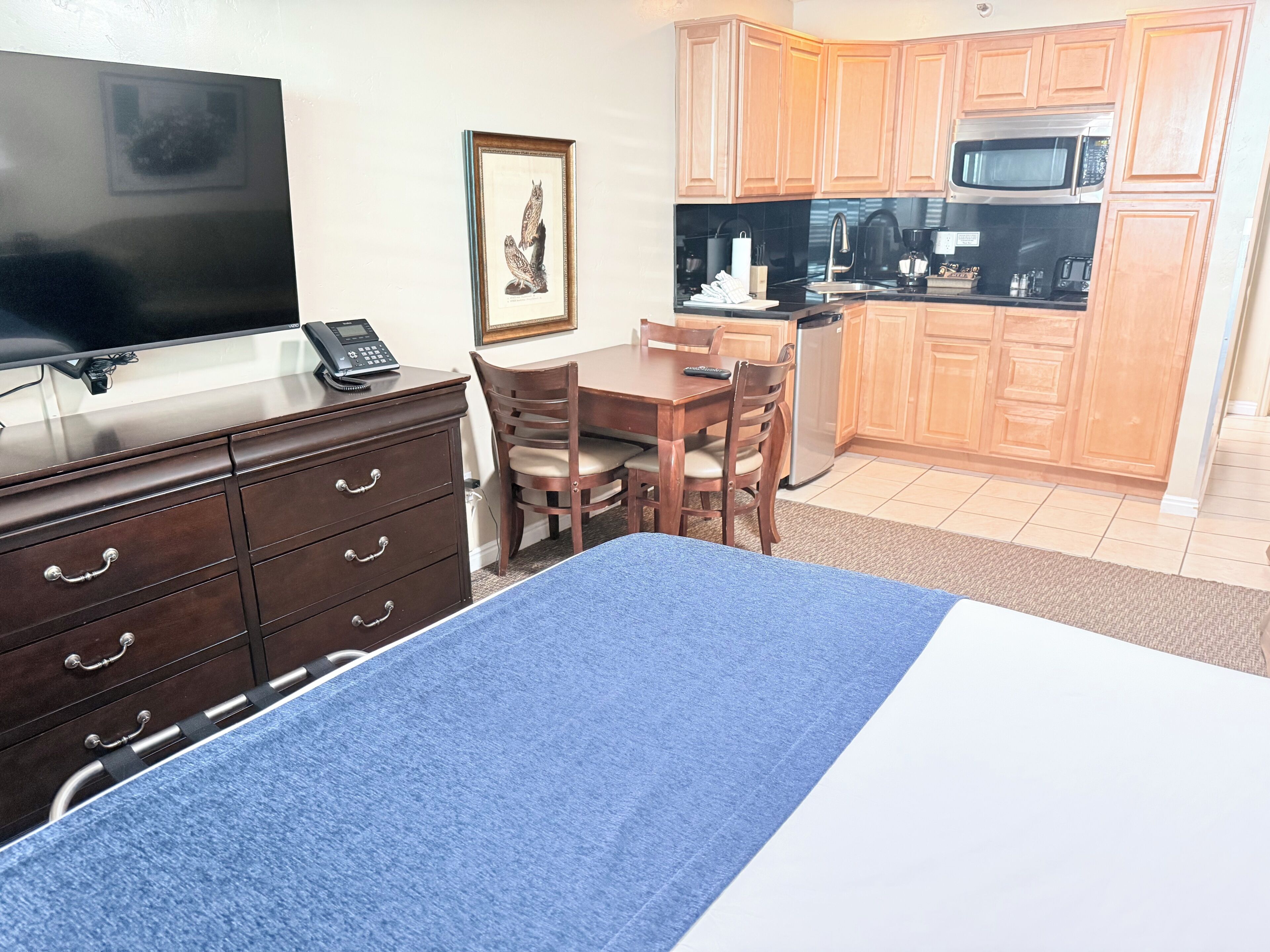 A hotel room with a bed, a desk, a chair, a television, a telephone, a framed picture, and a kitchenette.