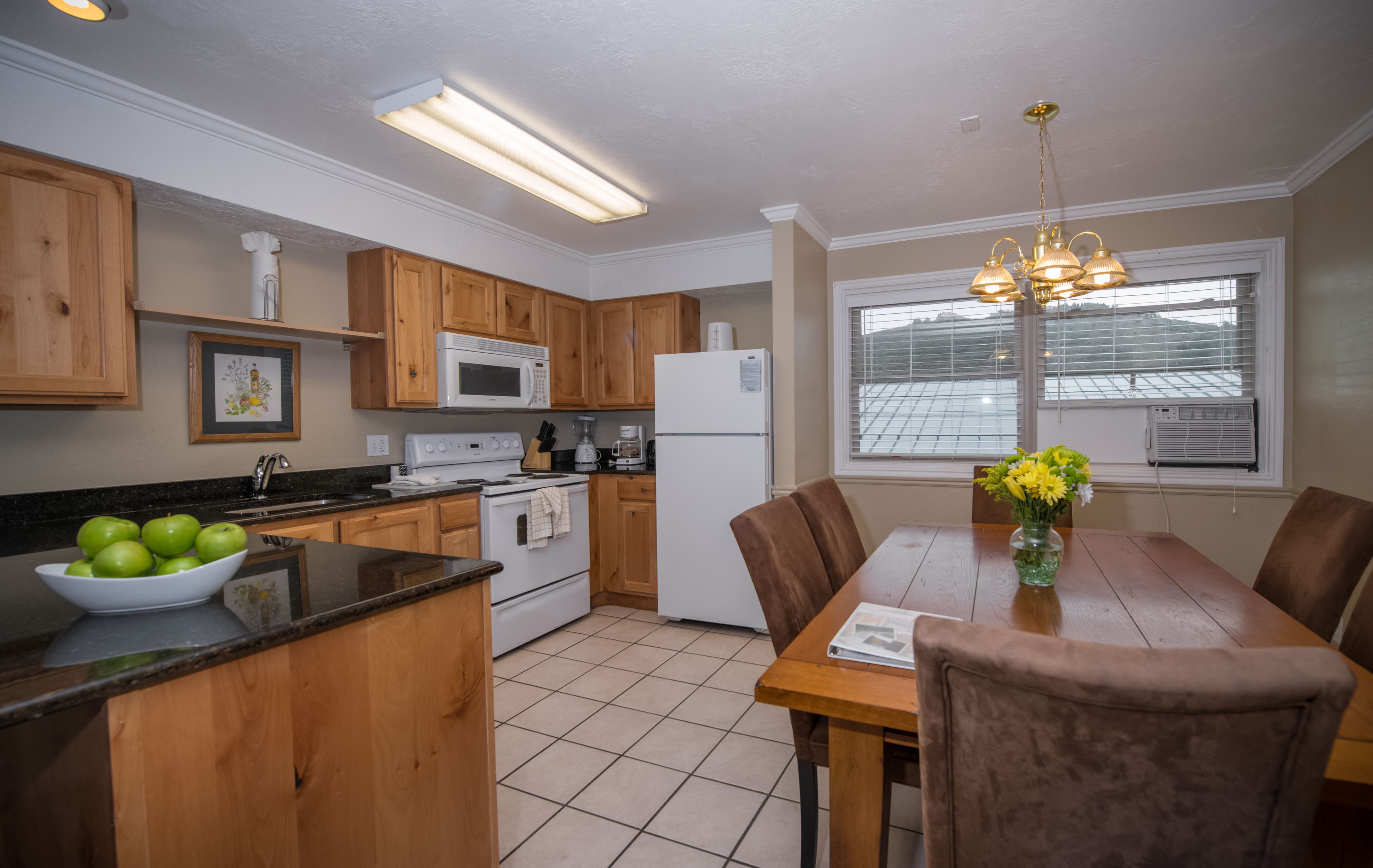 A kitchen with wooden cabinets, a microwave, a refrigerator, a dining table with chairs, and a window with blinds.