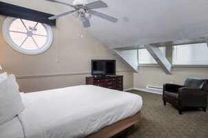 Un dormitorio con cama, televisor, silla, ventilador de techo y ventana.