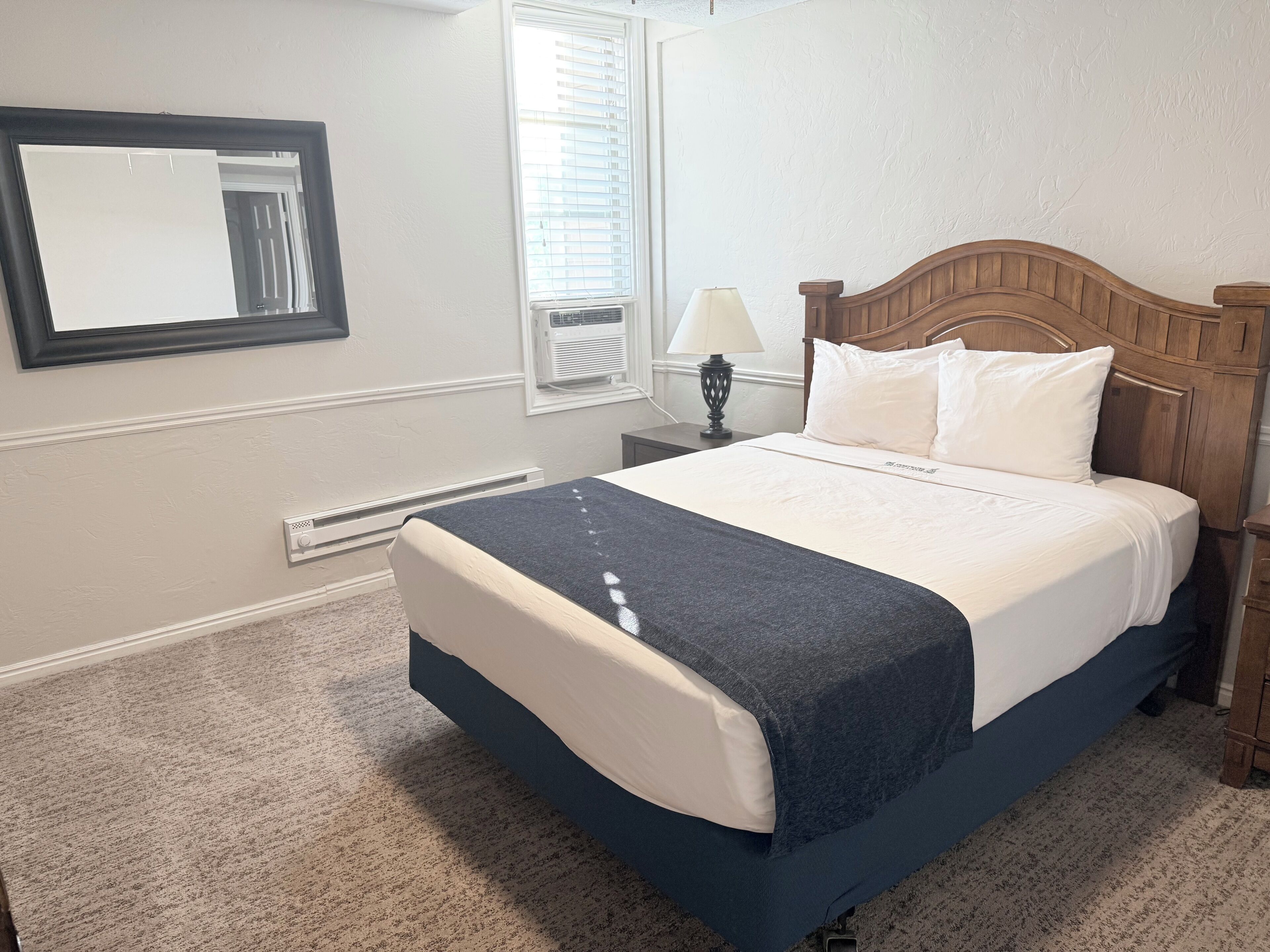 A neatly made bed with a wooden headboard, a bedside lamp, and a window with blinds.
