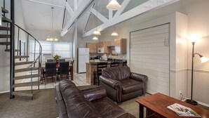 Amplia sala de estar con un sofá de cuero marrón, una mesa de centro de madera y una mesa de comedor con sillas. Se ve una cocina al fondo, con armarios de madera y un refrigerador blanco.