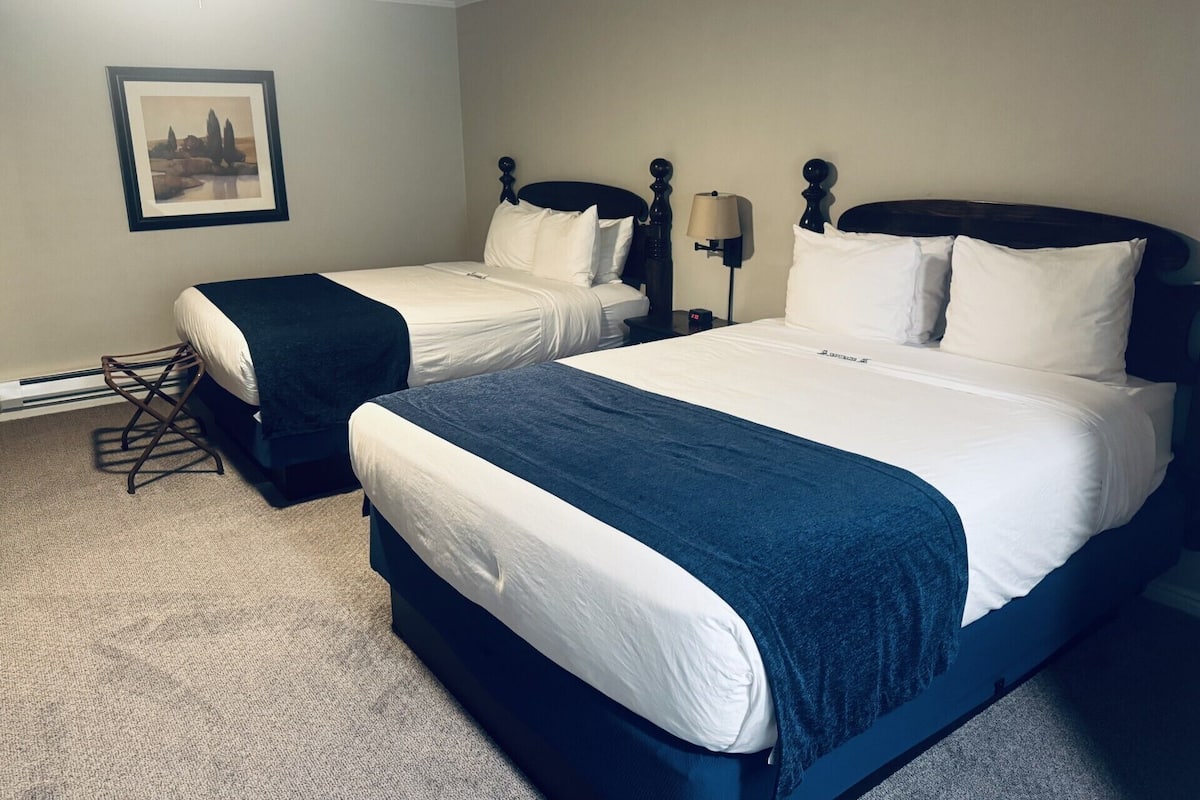 A hotel room with two beds, a nightstand, a lamp, a framed picture on the wall, and a carpeted floor.