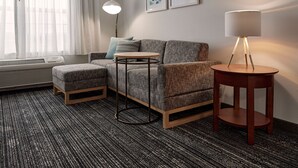 Ein modernes Hotelzimmer mit grauem Sofa, einem runden Holztisch, einer Stehlampe und Bildern an der Wand.