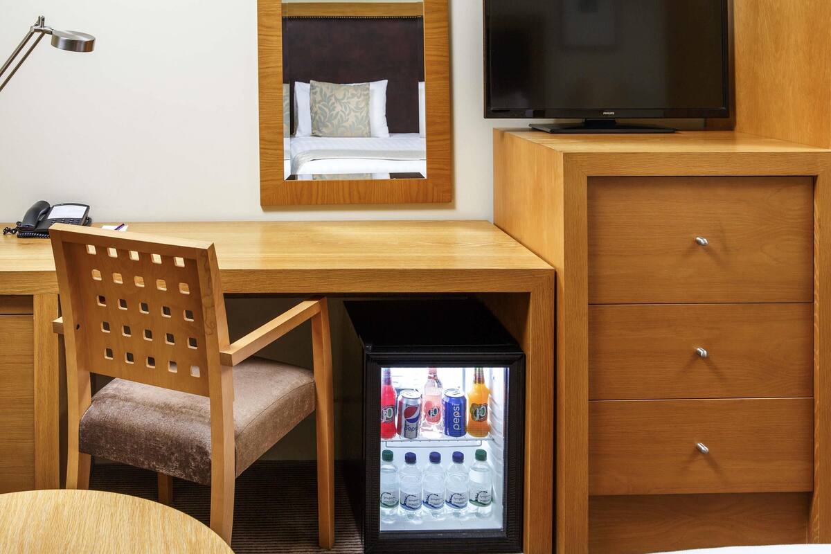 Una camera d'albergo con una scrivania in legno, una sedia, un televisore, un minibar e un letto.
