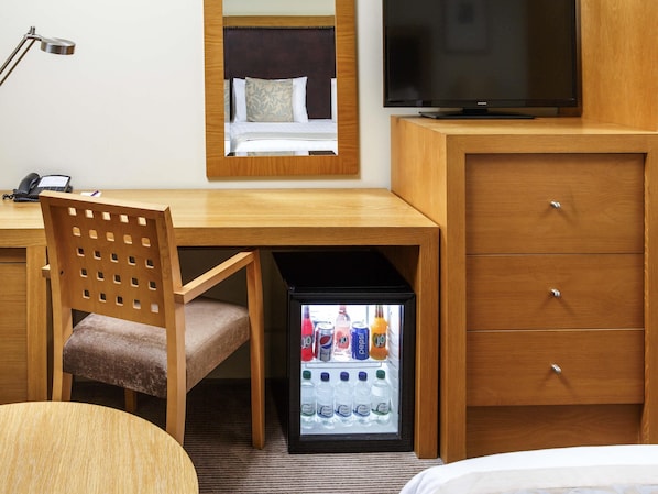 A hotel room with a wooden desk, a chair, a television, a minibar, and a bed.
