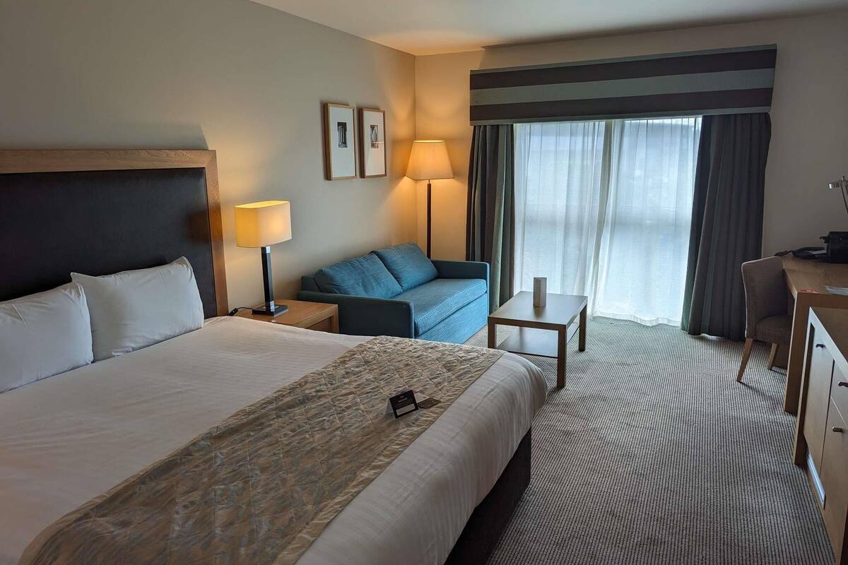 Una camera d'albergo con un letto, una scrivania, una sedia, un divano, una lampada e una finestra con tende.