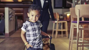 A child with a teddy bear and a suitcase in a hotel corridor with wooden chairs and tables.