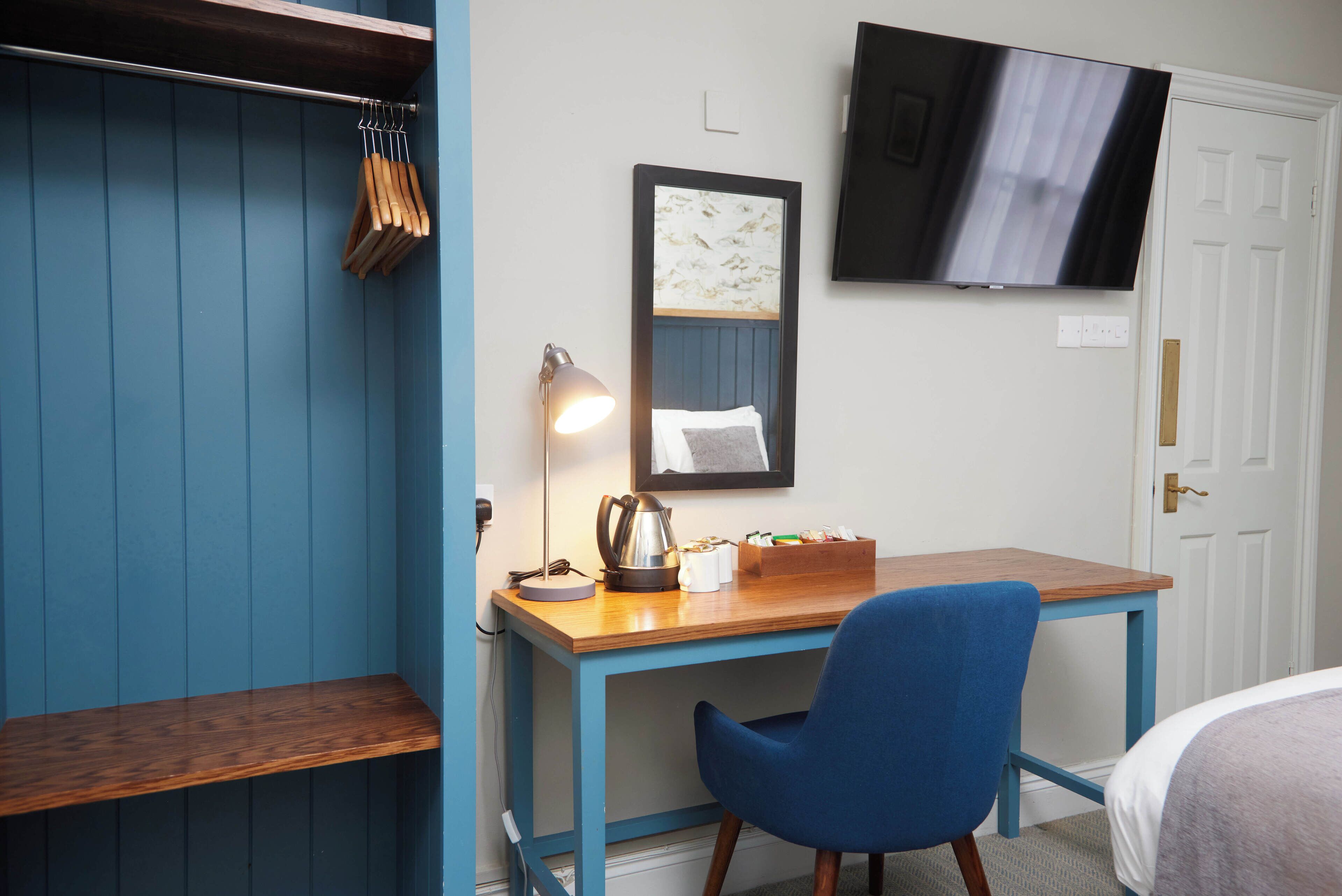 Una habitación con un escritorio de madera, una silla azul, un espejo, un televisor y un perchero.