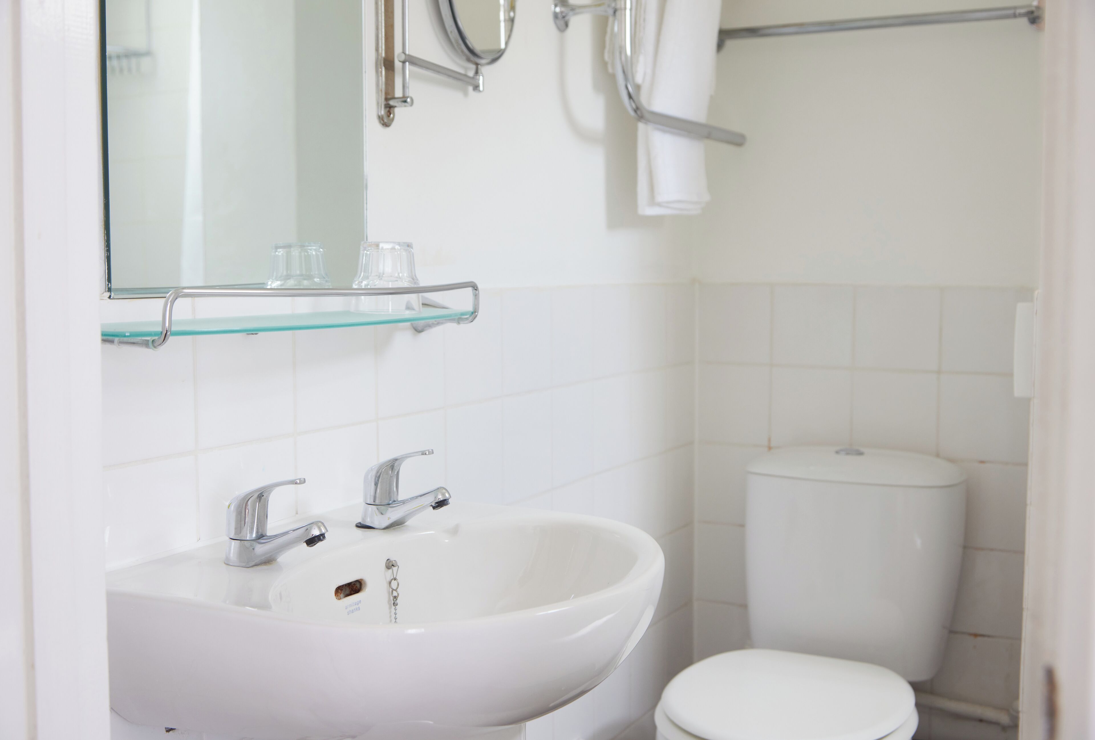 A bathroom with a white sink, two faucets, a glass shelf, a mirror, and a toilet.