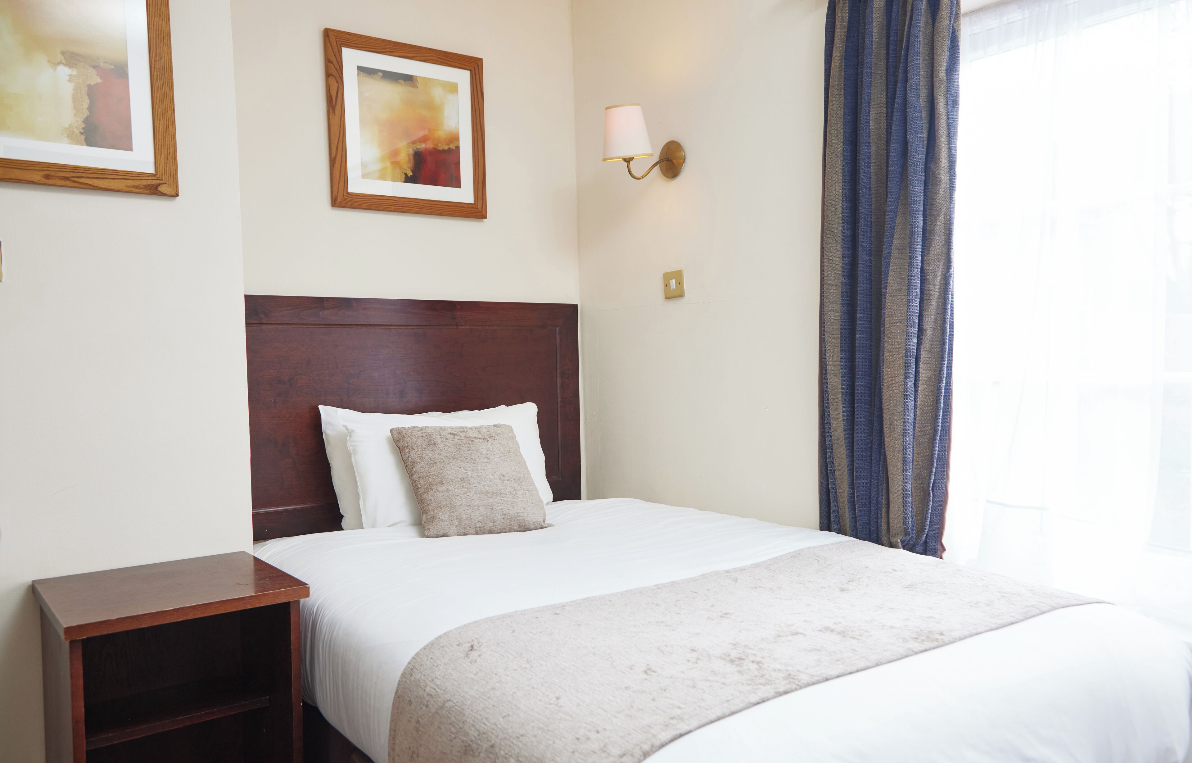 Une chambre d’hôtel avec un grand lit, une tête de lit en bois, une table de chevet, une lampe fixée au mur et une fenêtre avec des rideaux.