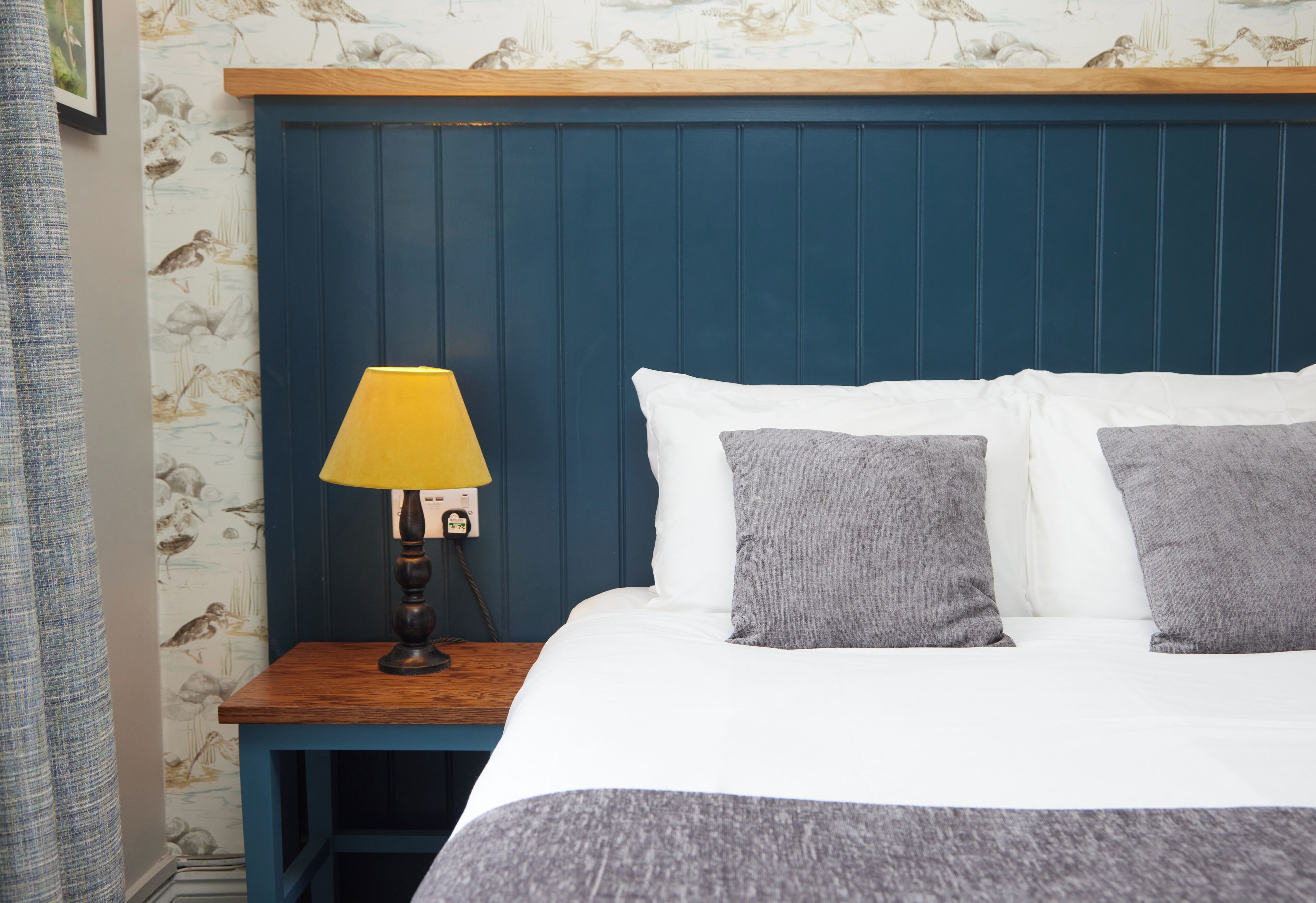 Ein Schlafzimmer mit einem blauen Kopfteil, einem Nachttisch aus Holz mit einer gelben Lampe sowie einem Bett mit weißer und grauer Bettwäsche.