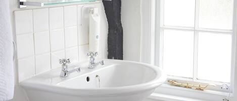 A bathroom with a white sink, two faucets, a glass shelf above the sink, a window with white frames, and a radiator below the window.