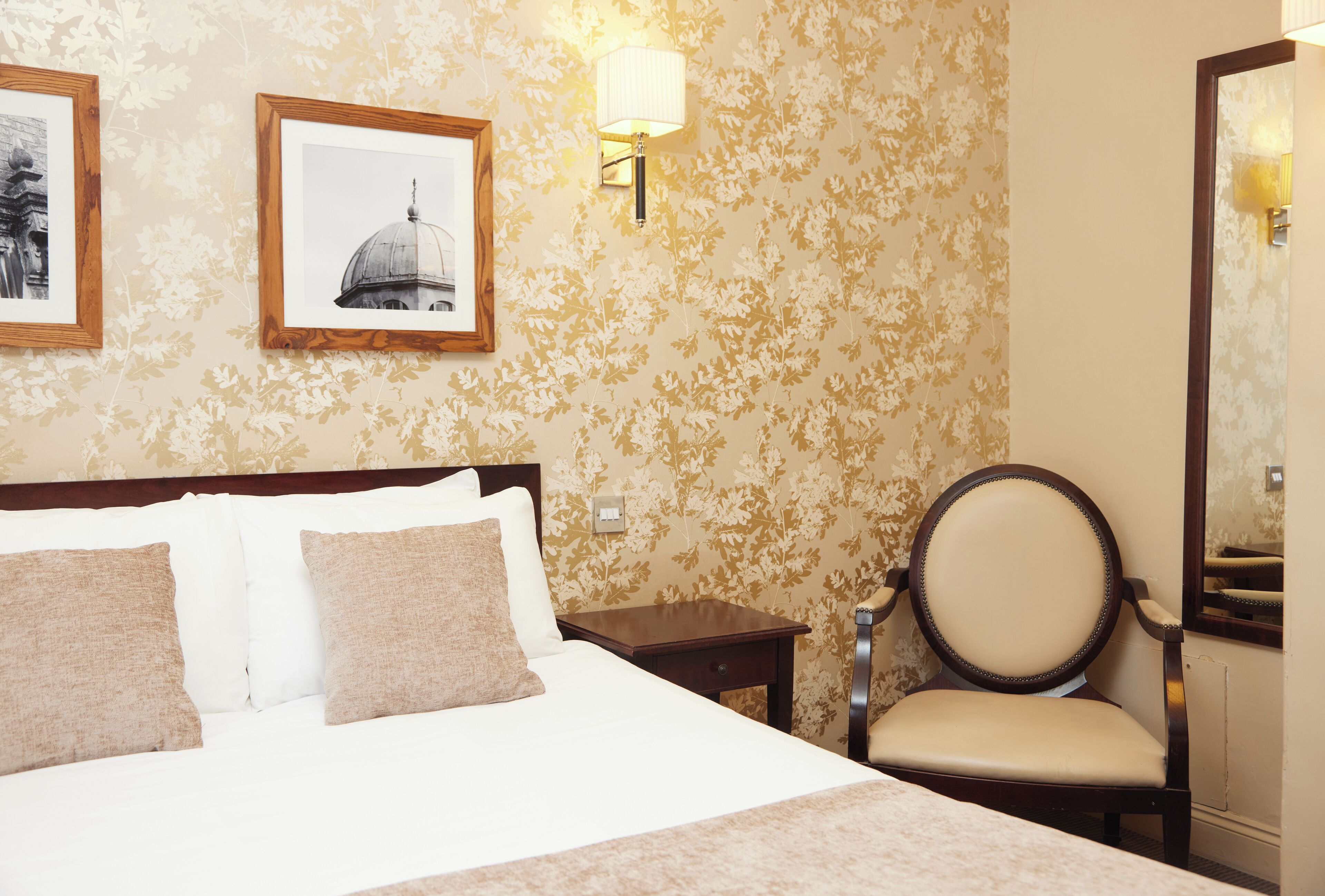 Habitación de hotel con una cama grande, dos cuadros en la pared, una mesita de noche, una silla y un espejo.