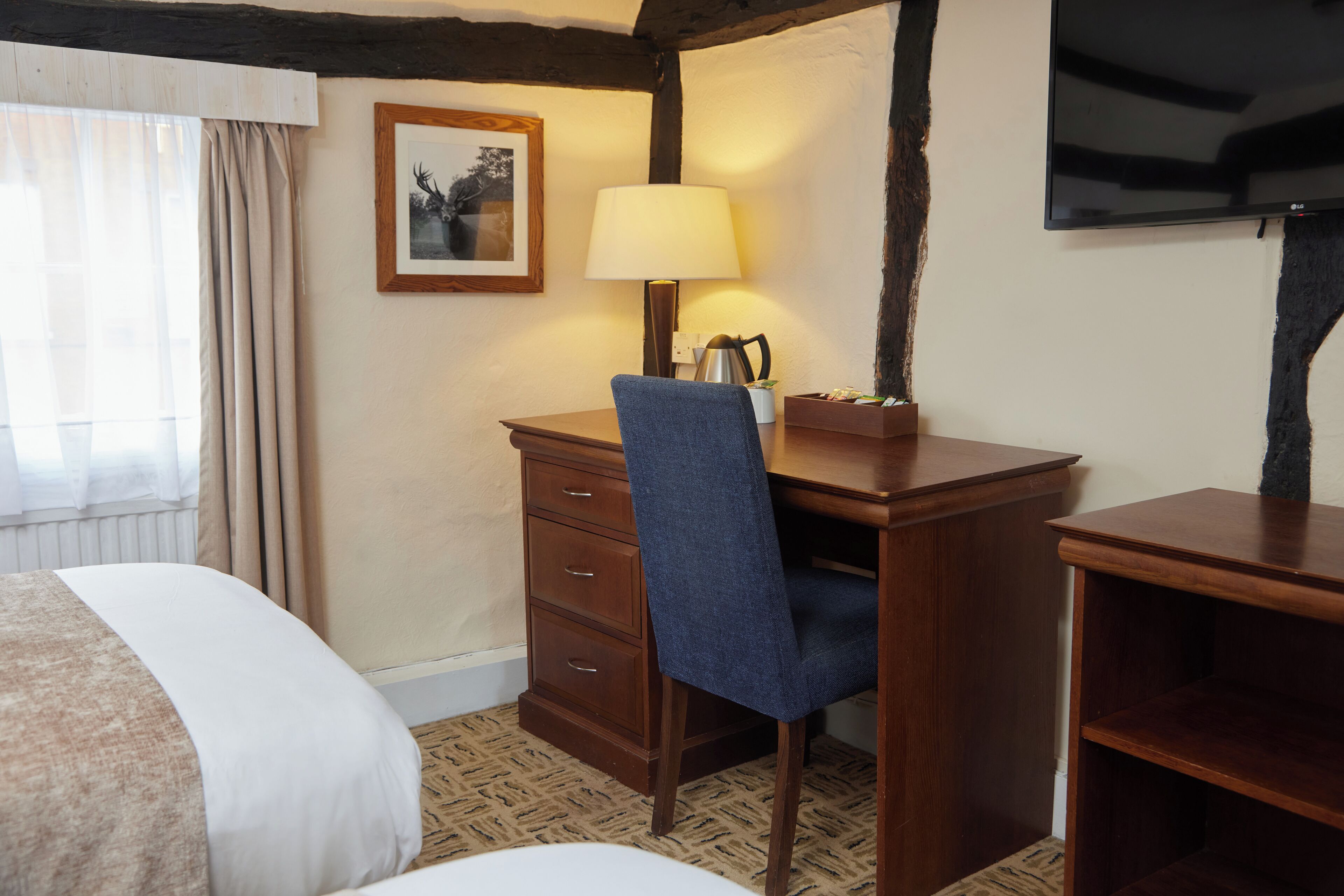 A hotel room with a bed, a desk with a chair, a lamp, a framed picture on the wall, and a television.