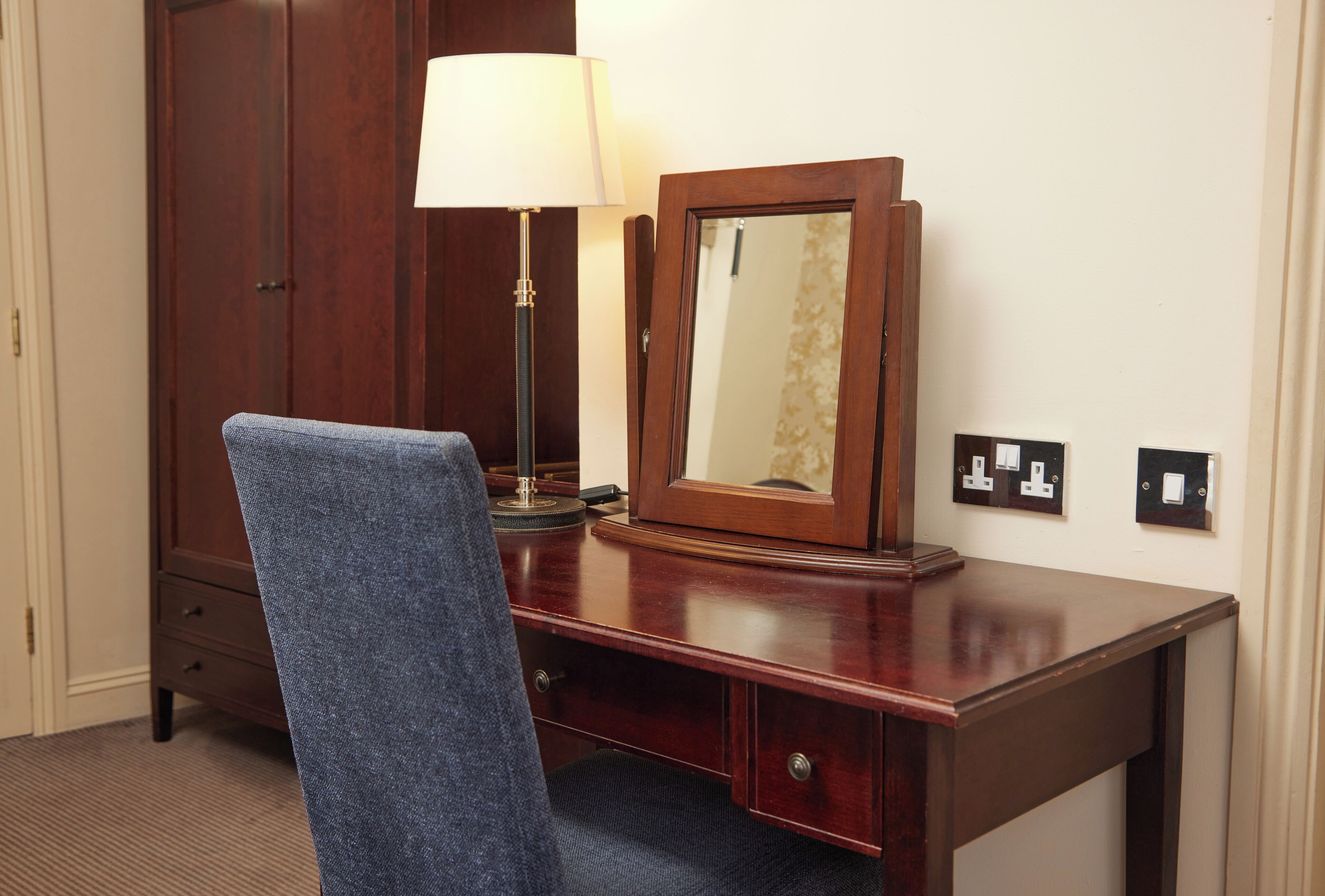 Un escritorio de madera con una lámpara, un espejo y una silla en una habitación de hotel.