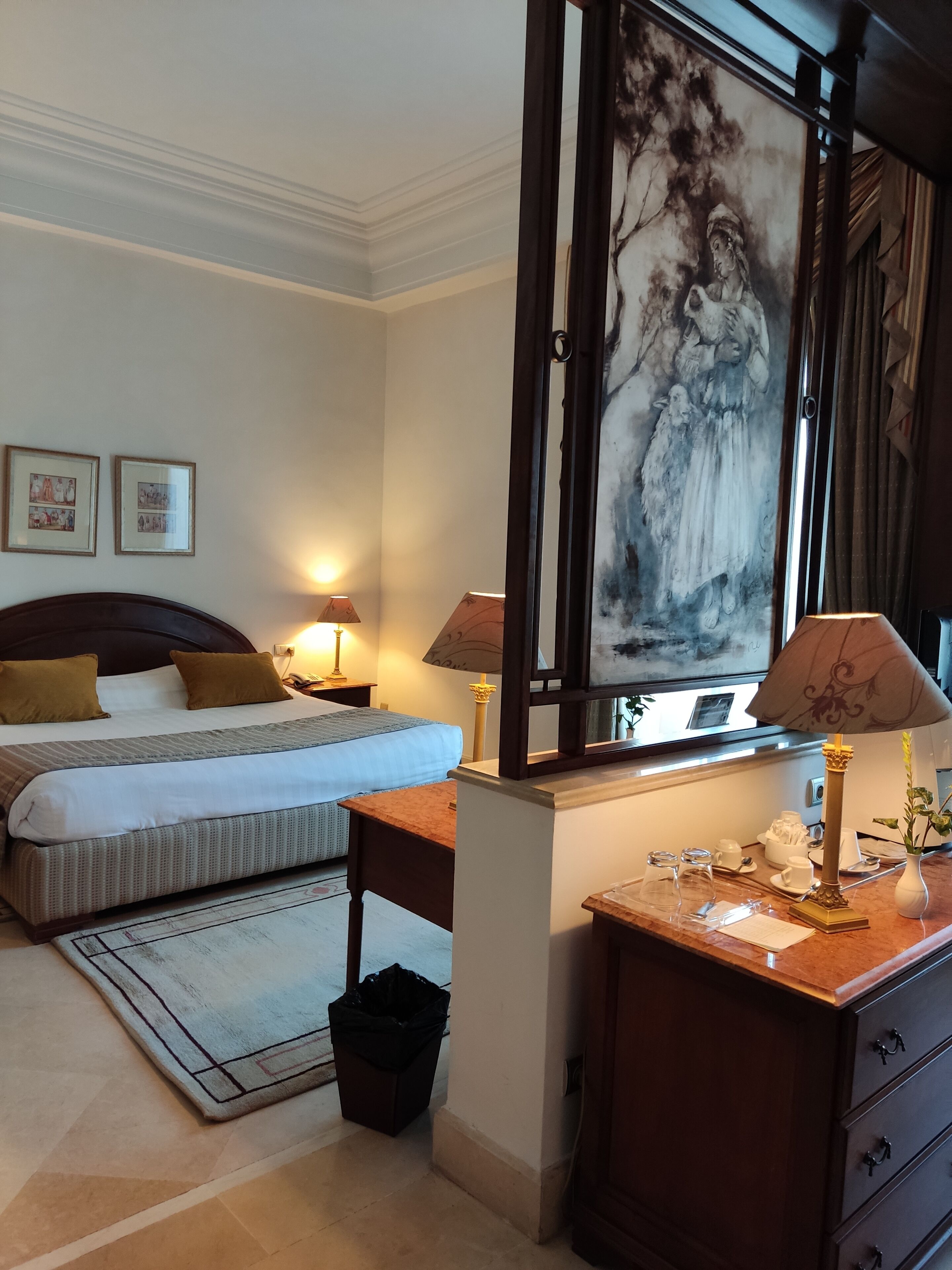 A hotel room with a large bed, two bedside tables with lamps, a desk with a lamp, a trash bin, and a large painting on the wall.