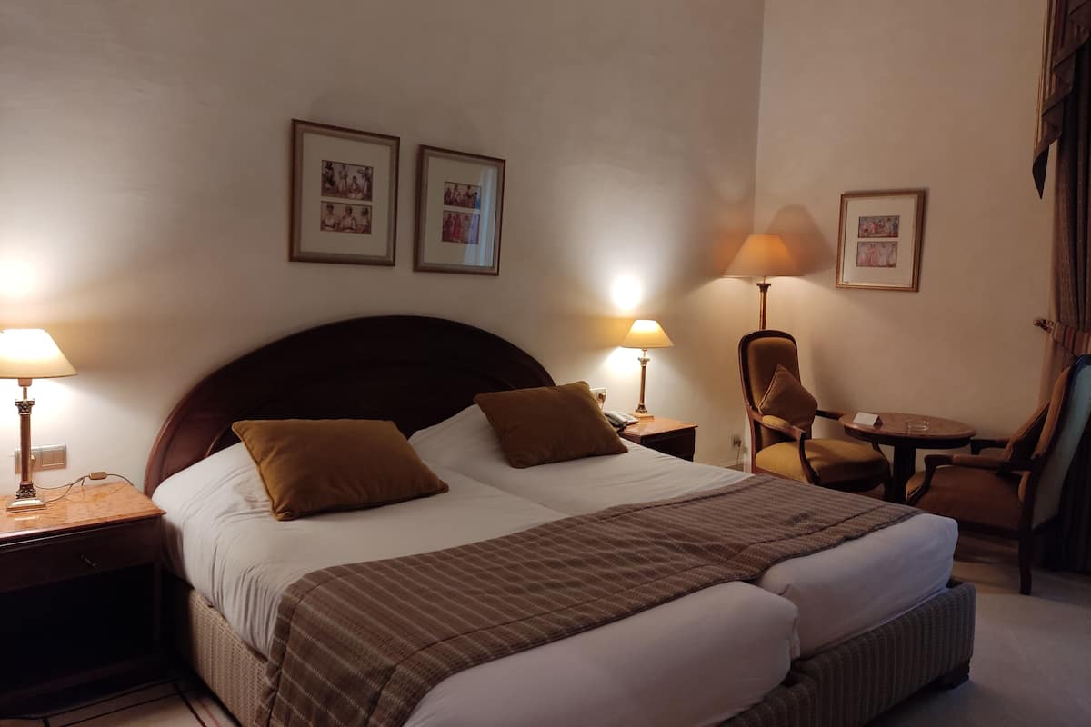 Quarto de hotel com uma cama grande, duas mesas de cabeceira com abajures, uma cadeira e quadros nas paredes.