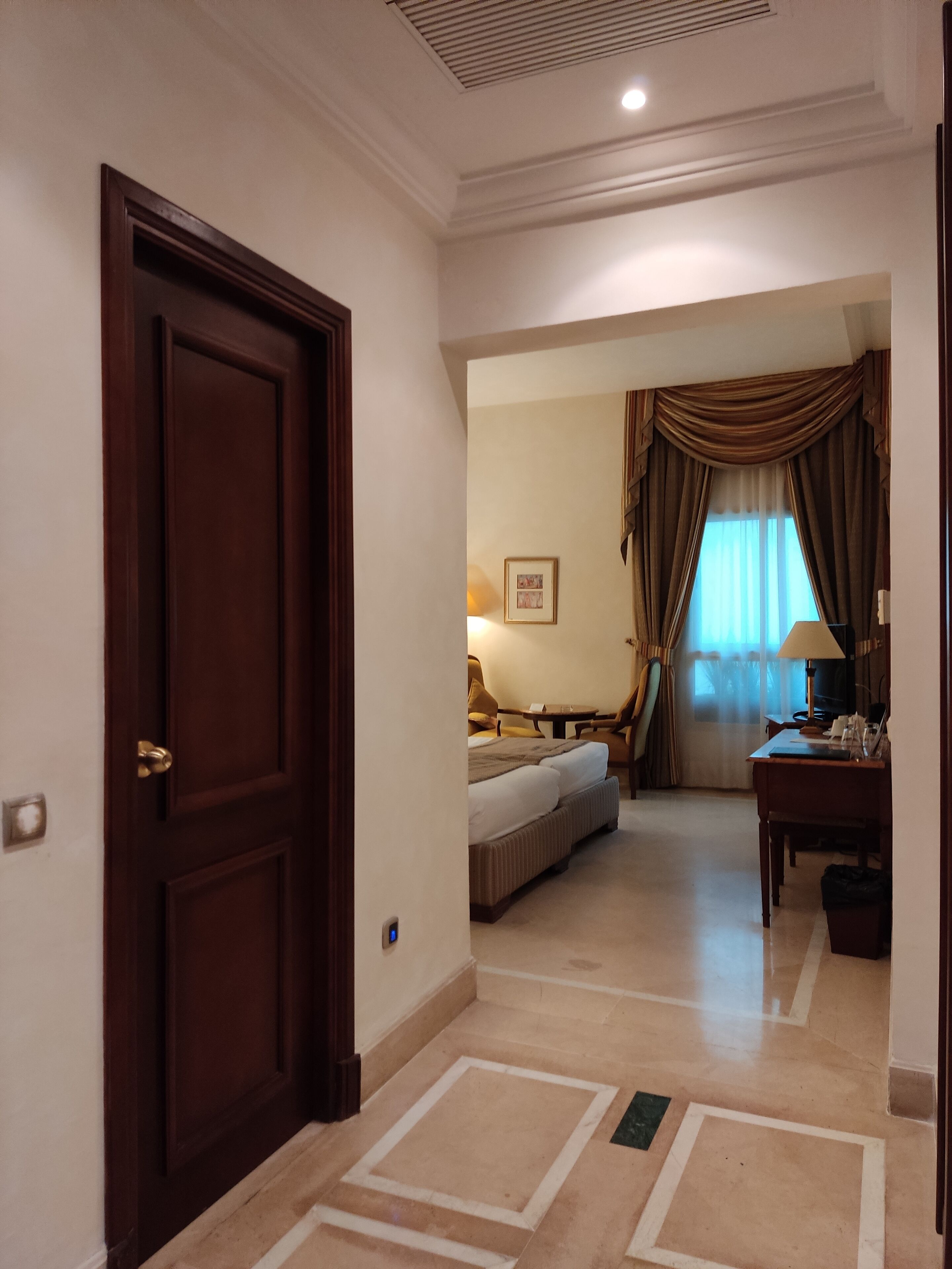 Habitación de hotel con una puerta de madera grande, una cama con cabecero, un escritorio con lámpara y una ventana con cortinas.