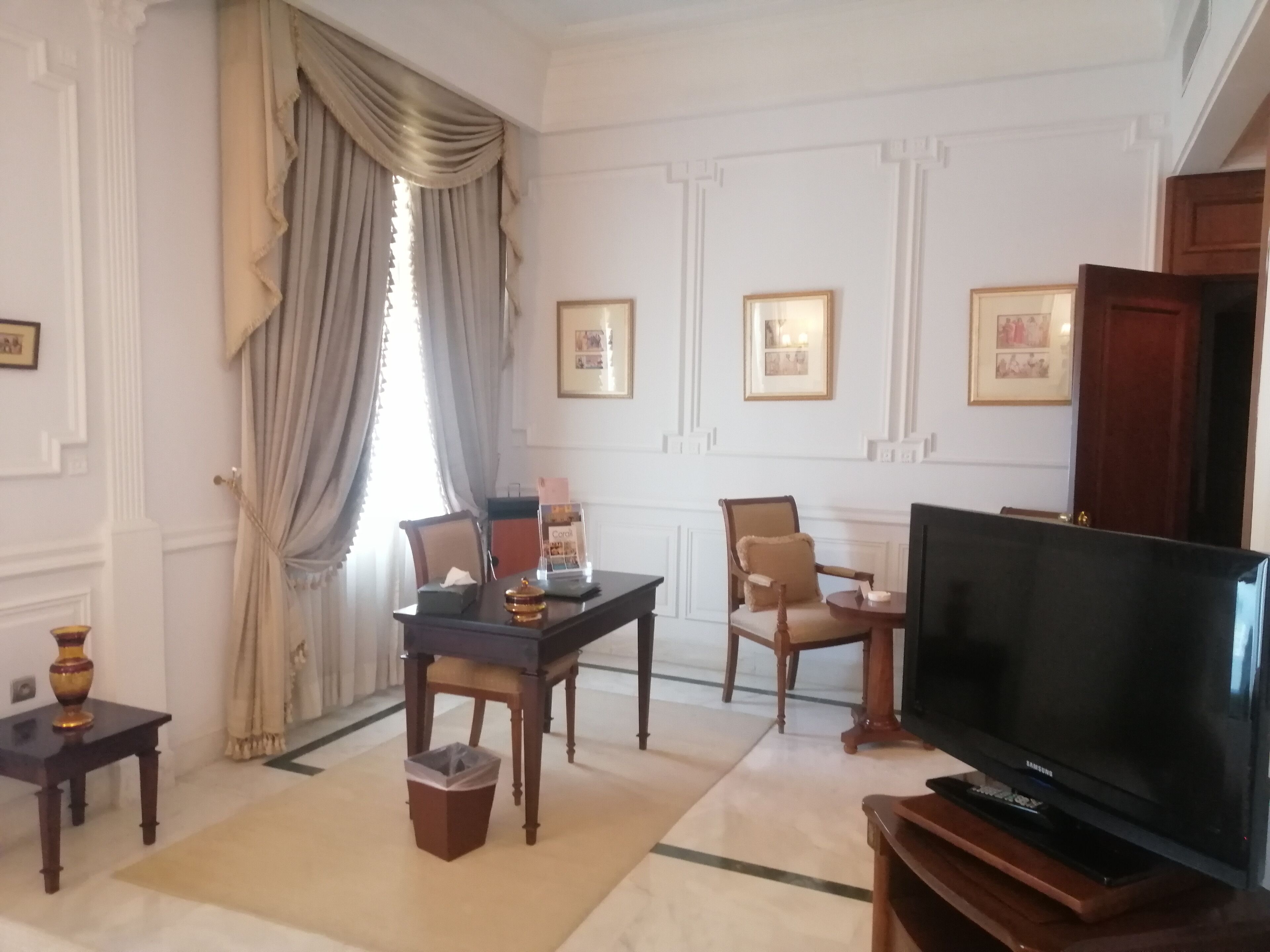 A hotel room with a large flat-screen TV, a dining table with chairs, a small trash bin, and a sofa.