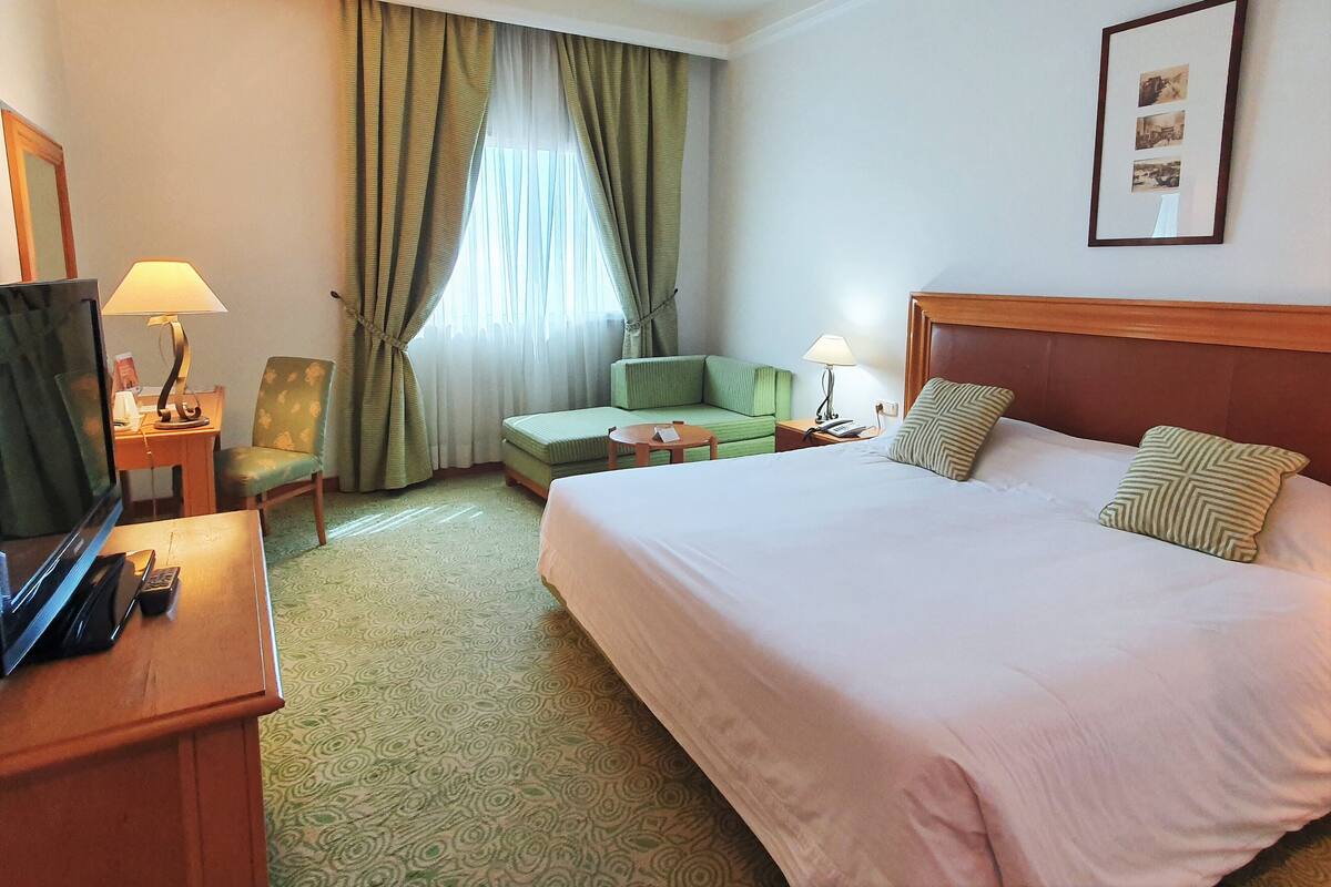 Quarto de hotel com uma cama grande, um sofá verde, uma escrivaninha com abajur e uma televisão.