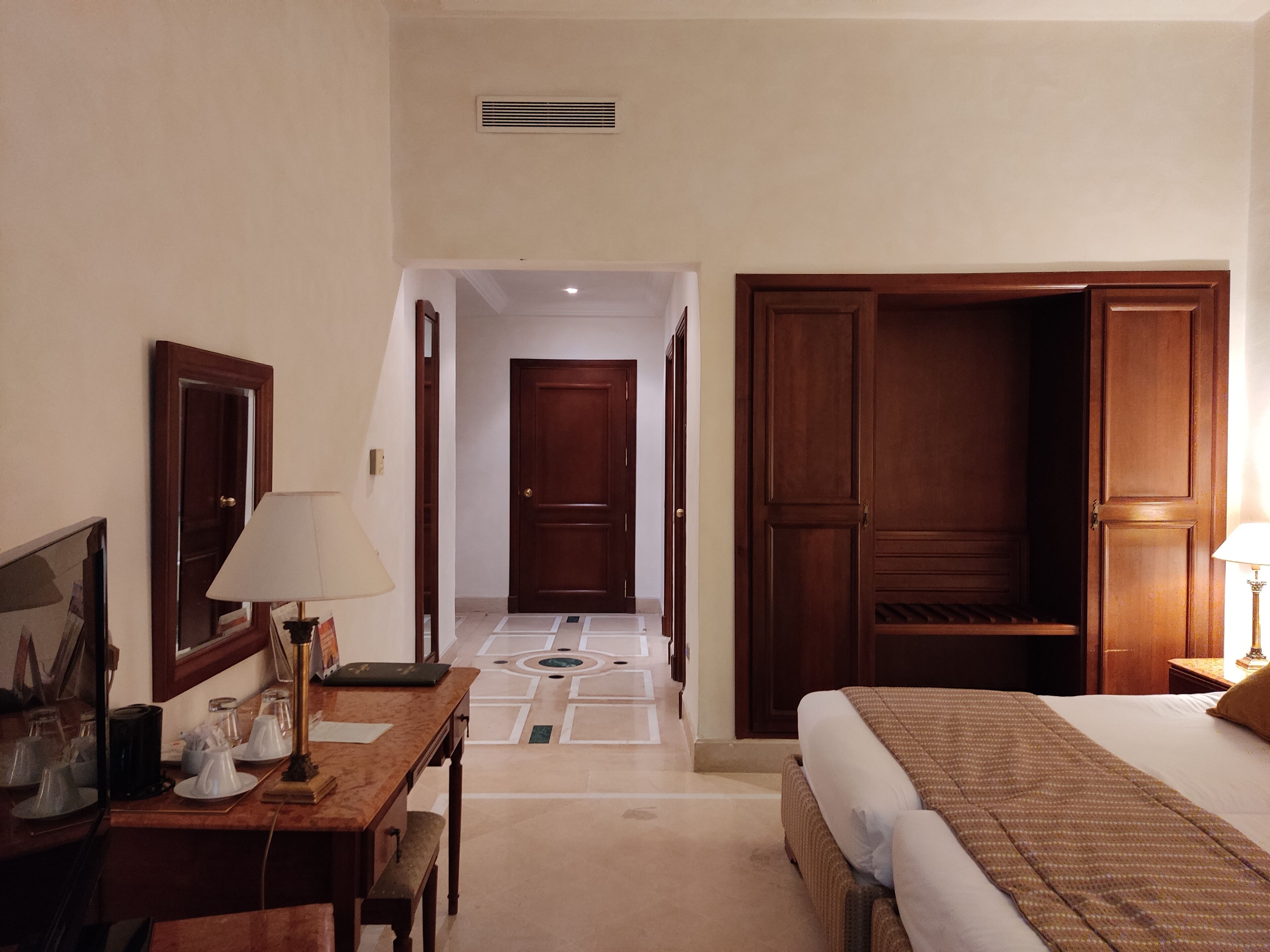 Habitación de hotel con cama, escritorio con lámpara, armario y puerta que da a otra habitación.