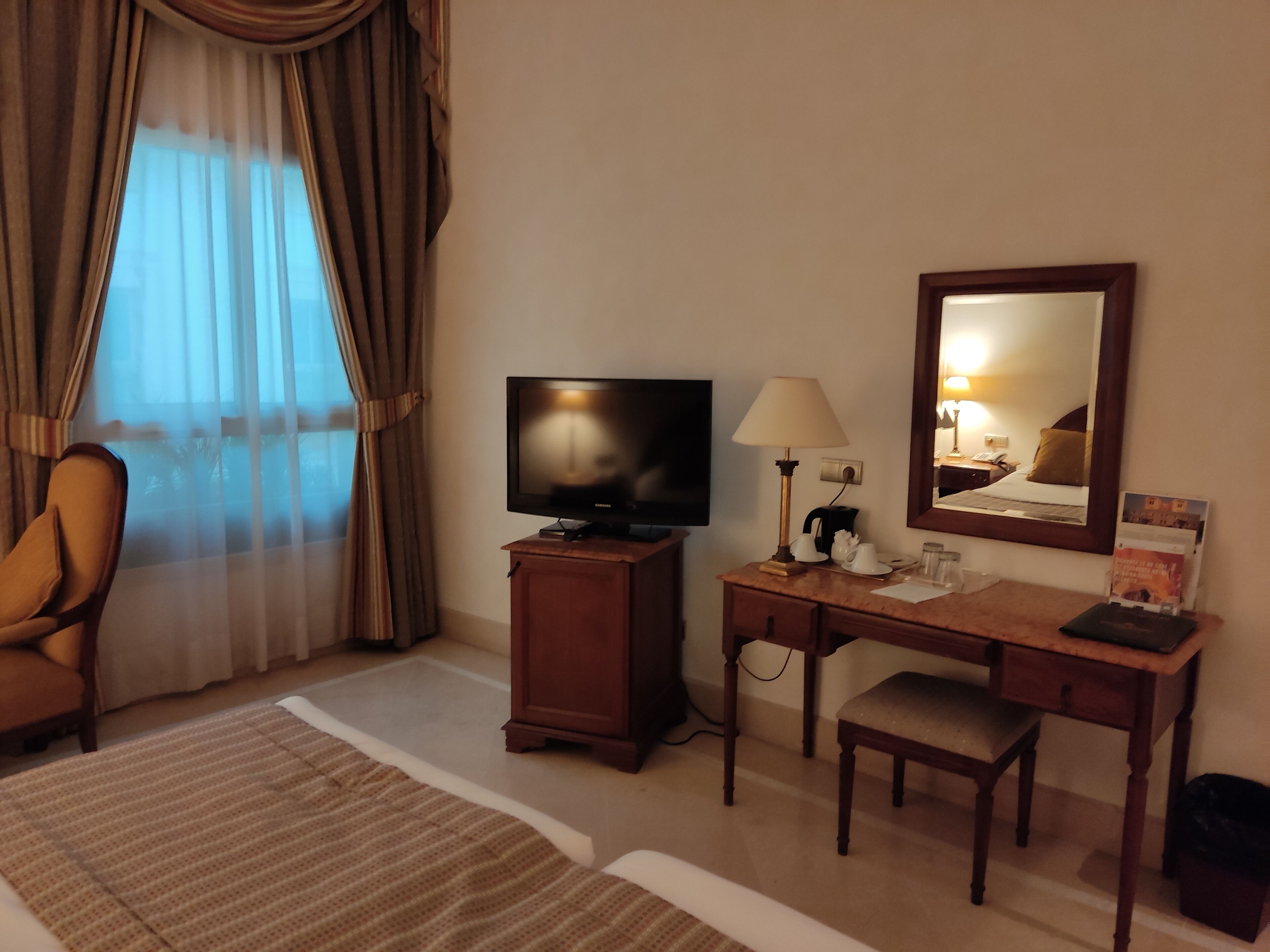 Habitación de hotel con televisión, escritorio, silla y un ventanal con cortinas.