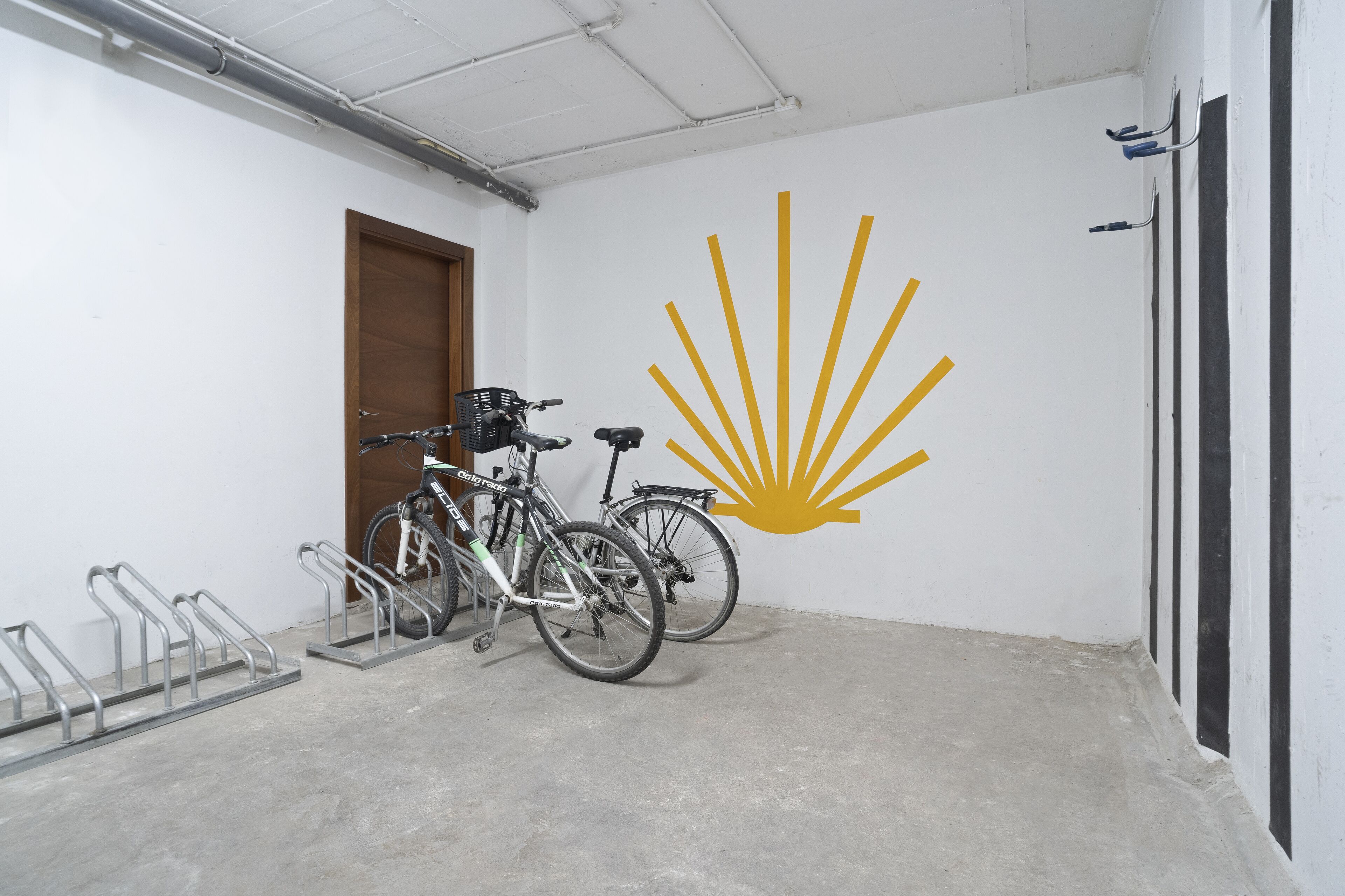 A room with a concrete floor, a wooden door, and a wall with a yellow sunburst logo. There are two bicycles parked against the wall.
