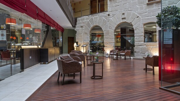 Amplio espacio interior con suelo de madera, sillas de mimbre y mesas, destacando una pared de piedra y puertas de cristal.