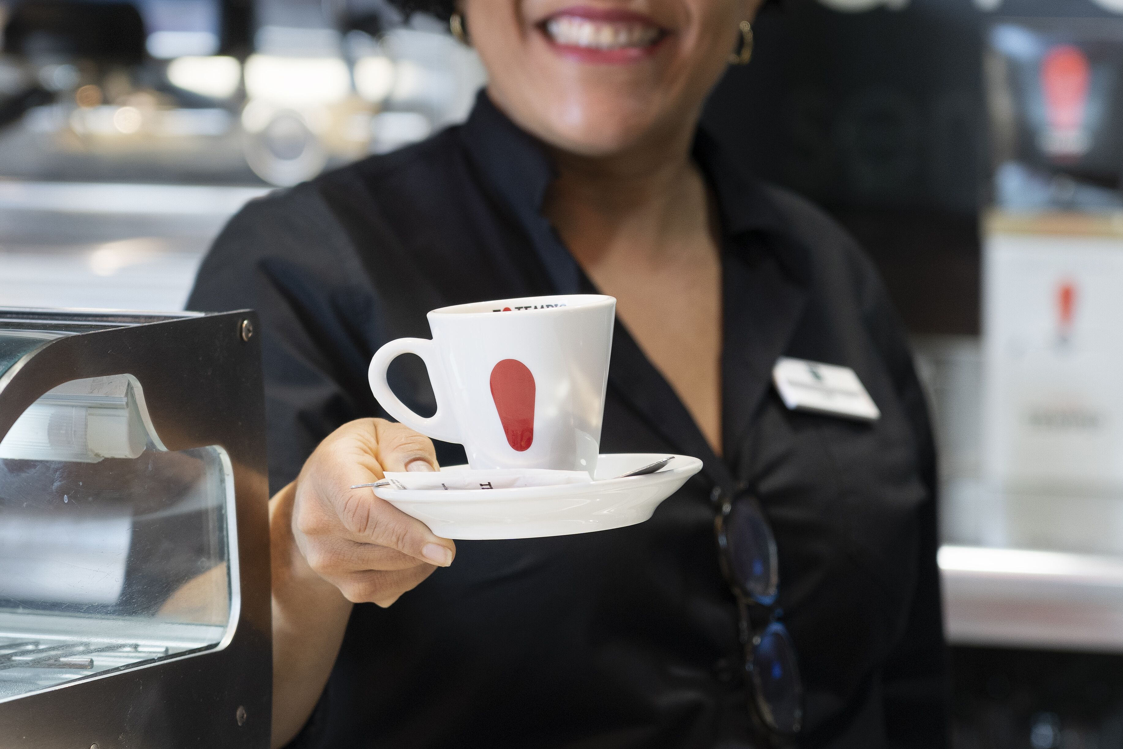 A person holding a white cup with a red heart design, wearing a black shirt with a name tag.
