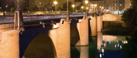 Un puente de piedra sobre un río, con su reflejo, rodeado de edificios y farolas.