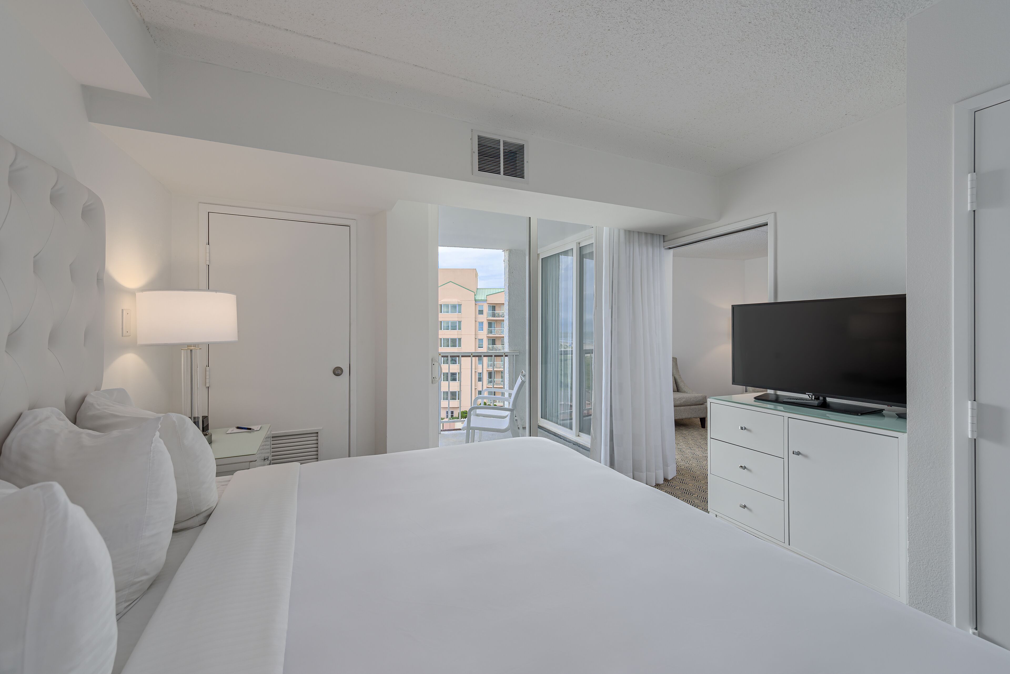 A hotel room with a large bed, a flat-screen TV, a desk with a lamp, and a view of the city through the window.