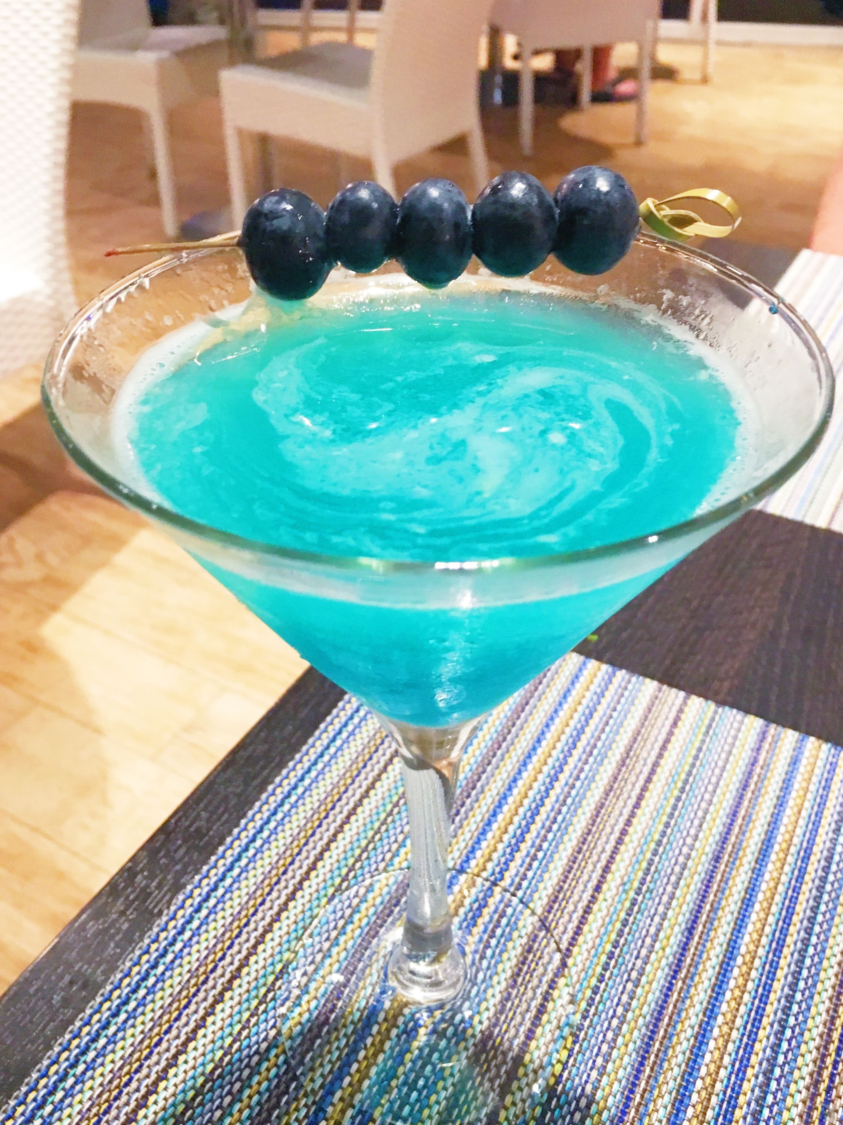 A cocktail in a martini glass with a blue liquid and blueberries on the rim.