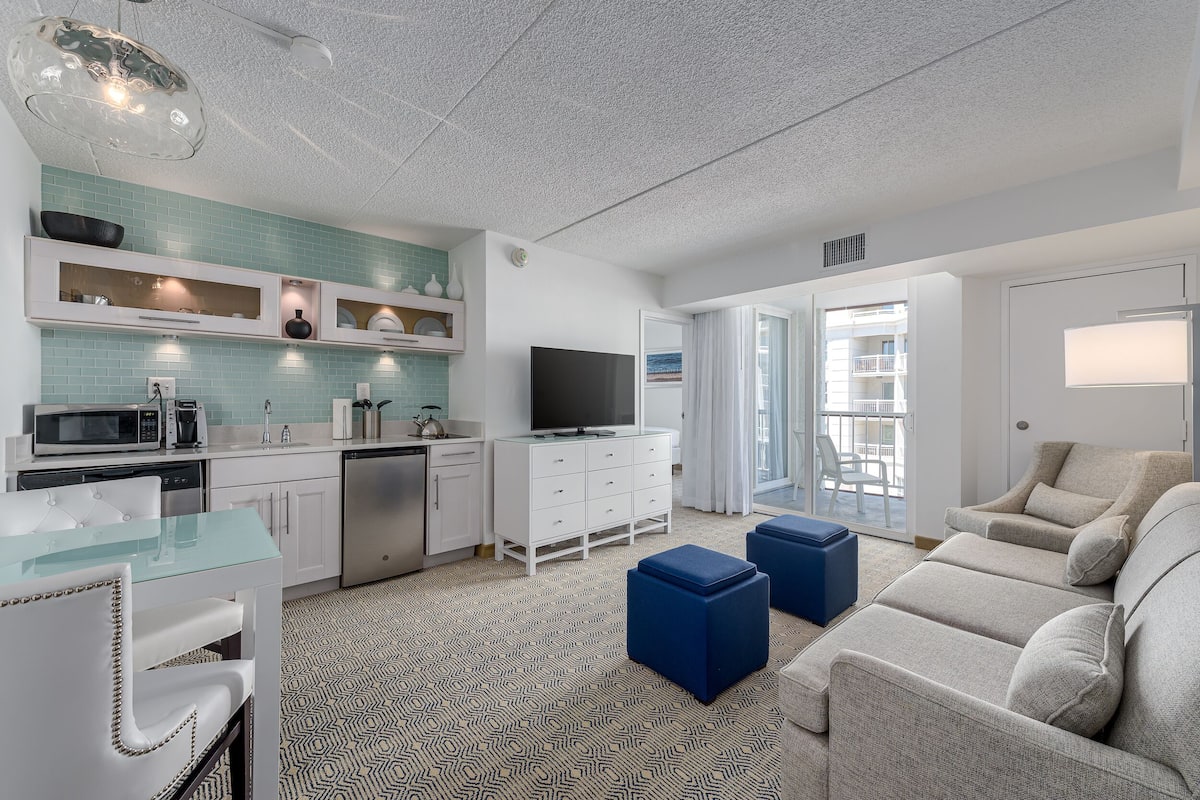 A modern living room with a sofa, ottomans, a TV, and a kitchen area with a microwave and cabinets.