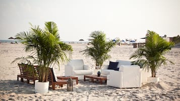 Un salón frente a la playa con sofás y mesas blancas, rodeado de palmeras en macetas.