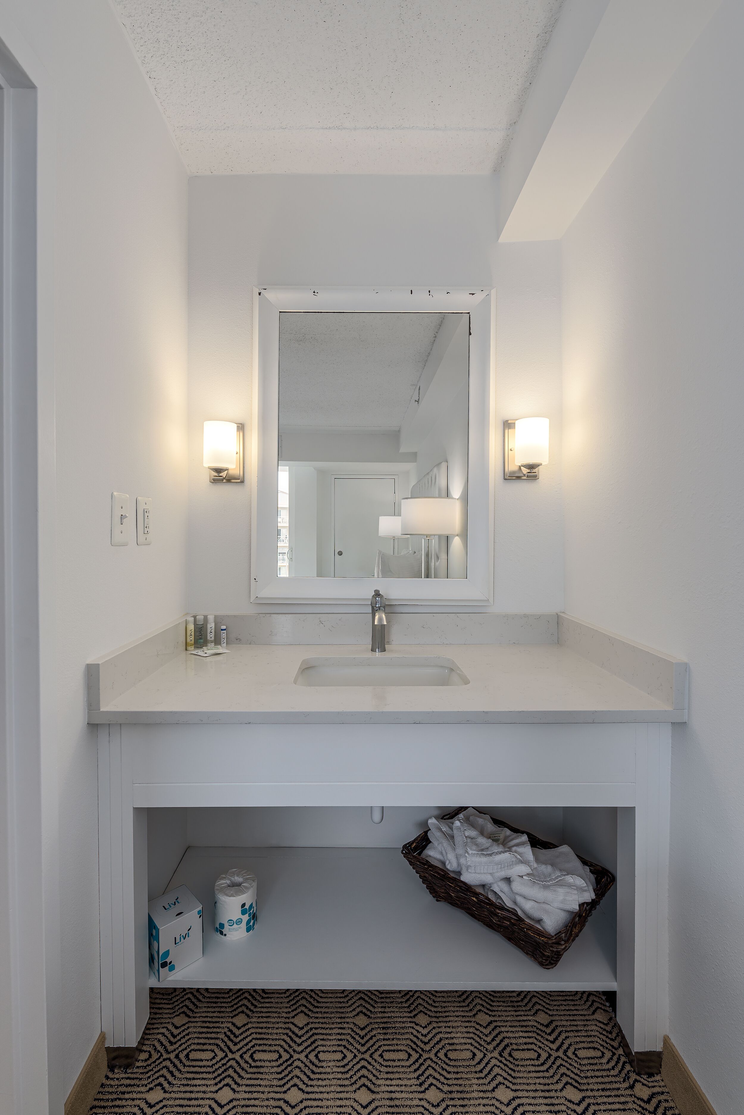 Un baño con un lavamanos blanco, espejo y dos luces empotradas en la pared. Hay una repisa debajo del lavamanos con una cesta y artículos de aseo.