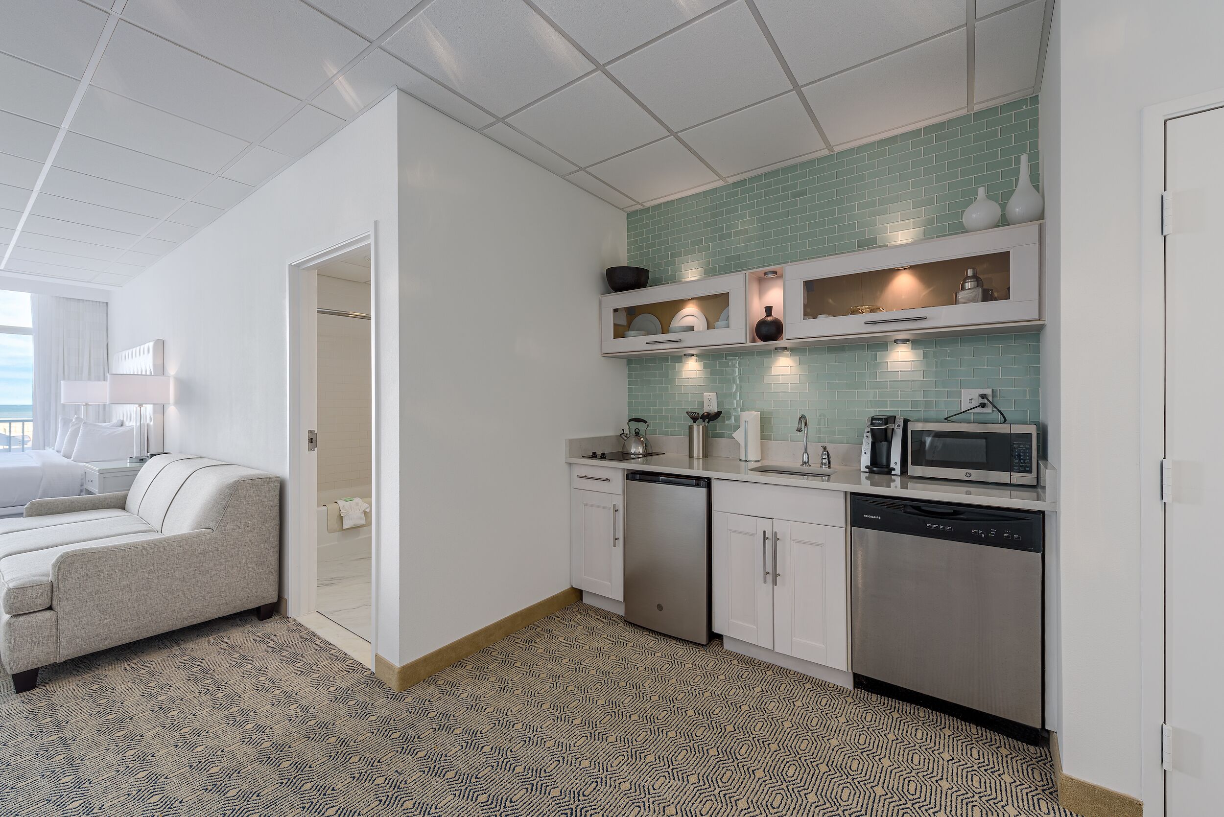 Una cocina moderna con armarios blancos, lavavajillas, microondas y un lavabo. Hay una alfombra estampada en el suelo y un sofá de color claro en la zona de estar contigua.