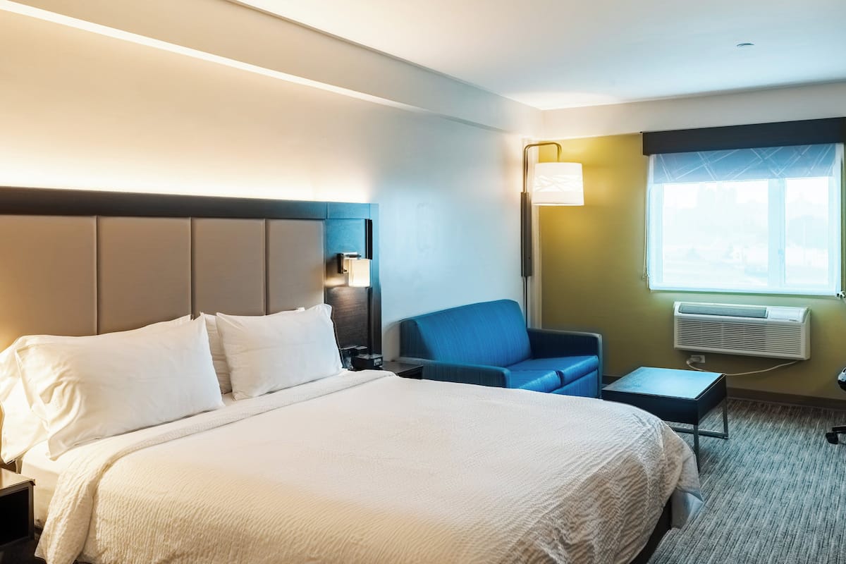 A hotel room with a large bed, a blue armchair, a small table, and a window with an air conditioner.