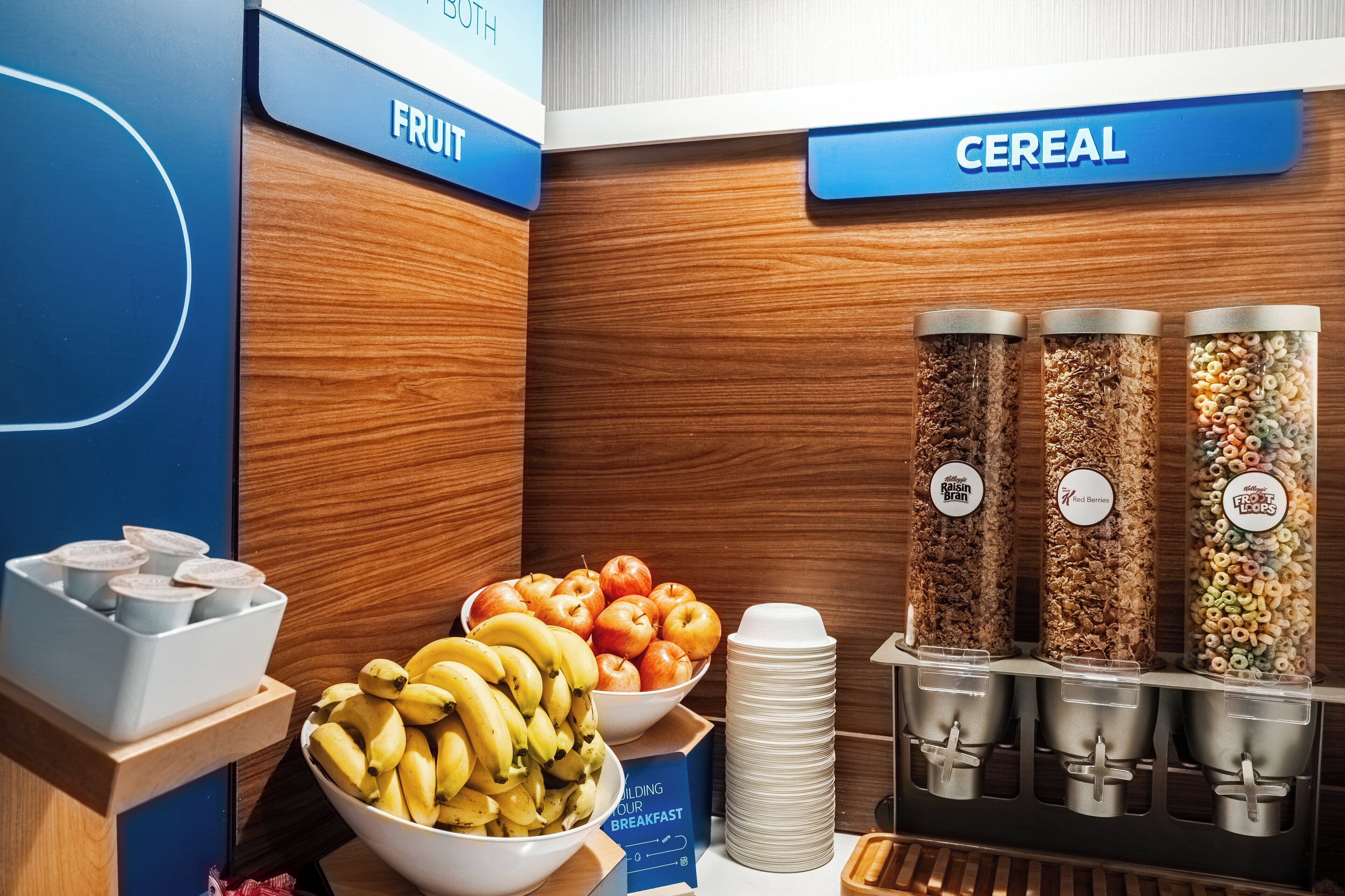 Un bufé de desayuno con secciones identificadas para fruta y cereales, incluyendo plátanos, manzanas y varias opciones de cereales.