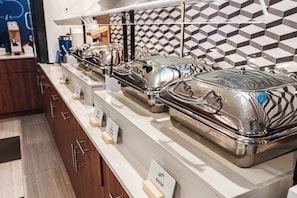 A buffet setup with multiple silver serving dishes labeled with food items. - Holiday Inn Express Maspeth by IHG (Maspeth)
