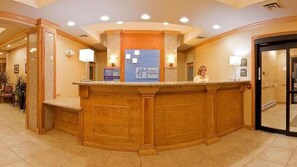A reception desk with a wooden finish, a blue sign with 'H' logo, and a woman standing behind it. - Holiday Inn Express Hotel & Suites Detroit - Utica by IHG (Utica)