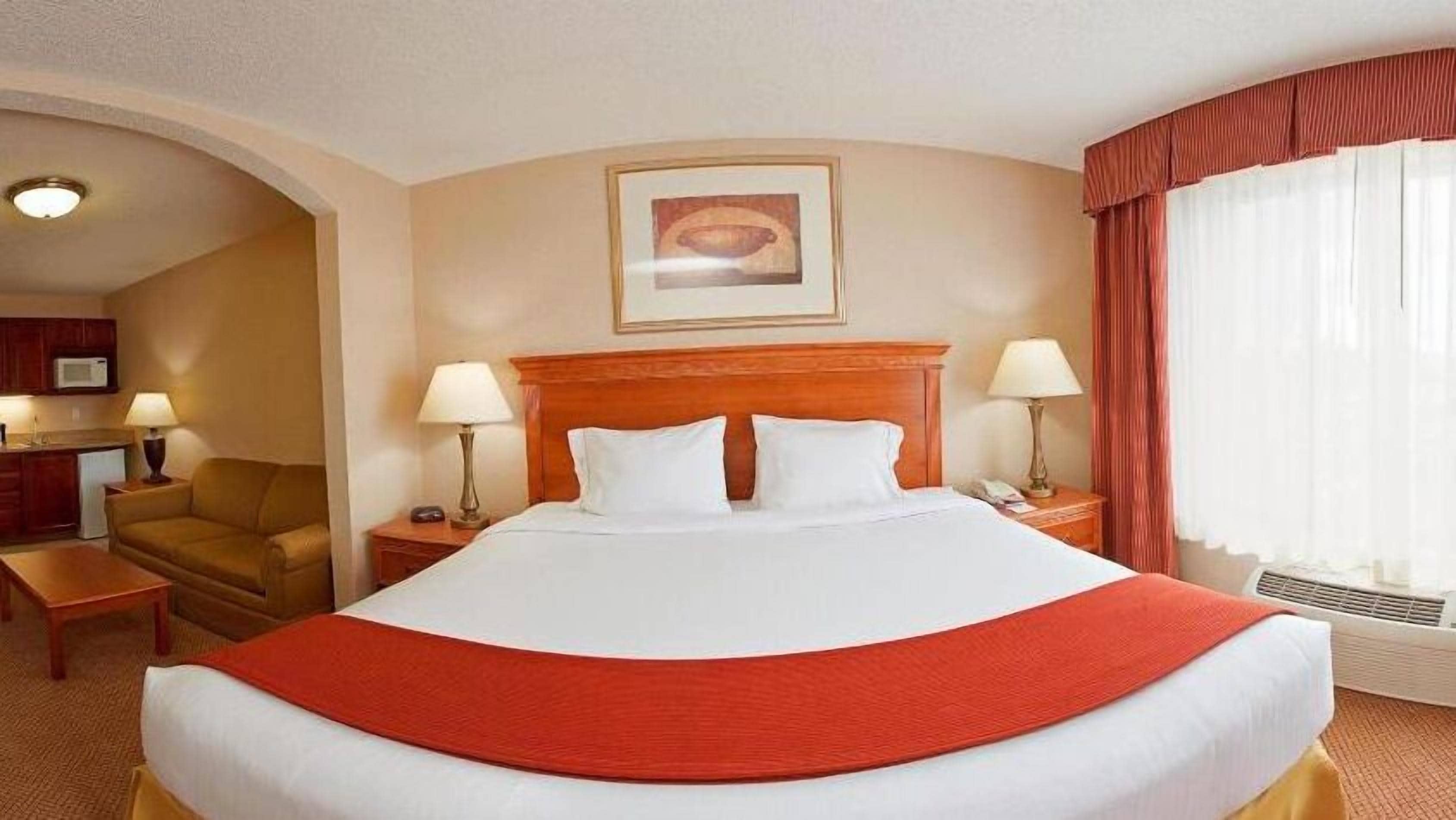 Una habitación de hotel con una cama grande, dos mesitas de noche con lámparas, un cabecero de madera, un cuadro en la pared y una ventana con cortinas.