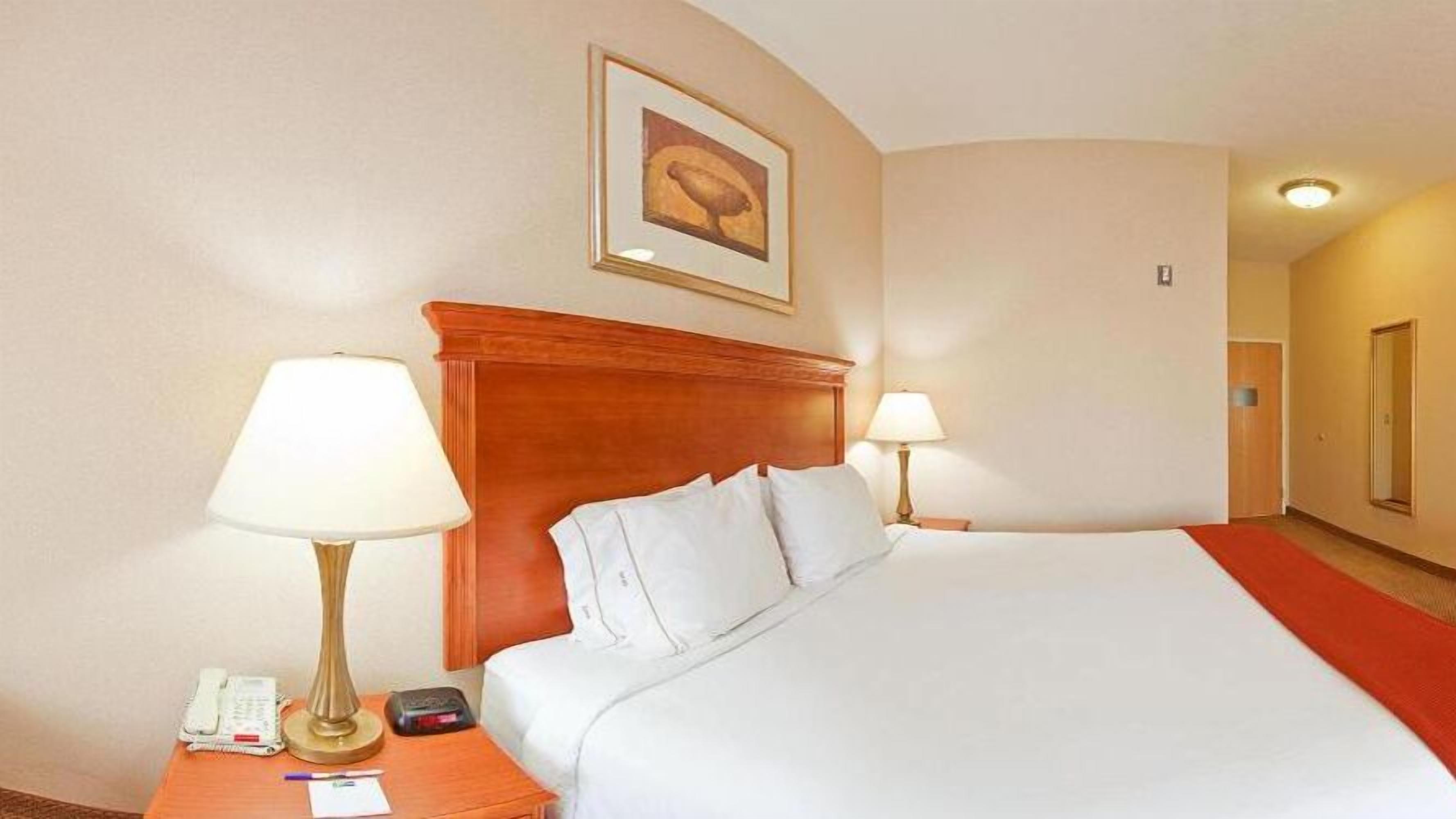 A hotel room with a large bed, two bedside lamps, a wooden headboard, a framed picture on the wall, and a telephone on the nightstand.