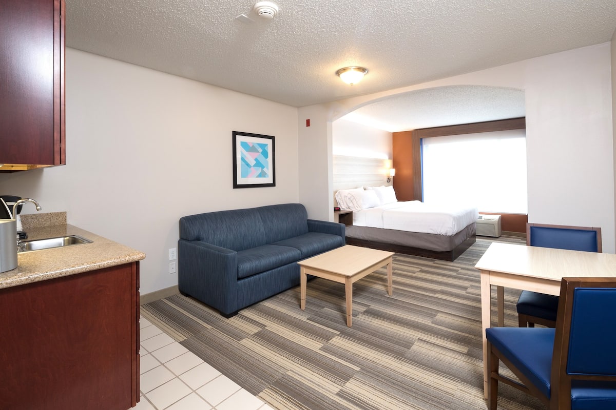A hotel room with a kitchenette, a sofa, a bed, a desk, and a window with blinds.