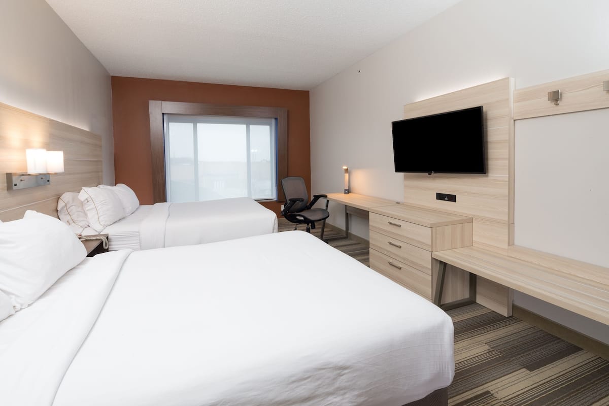A hotel room with two beds, a desk, a chair, a flat-screen TV, and a large window.