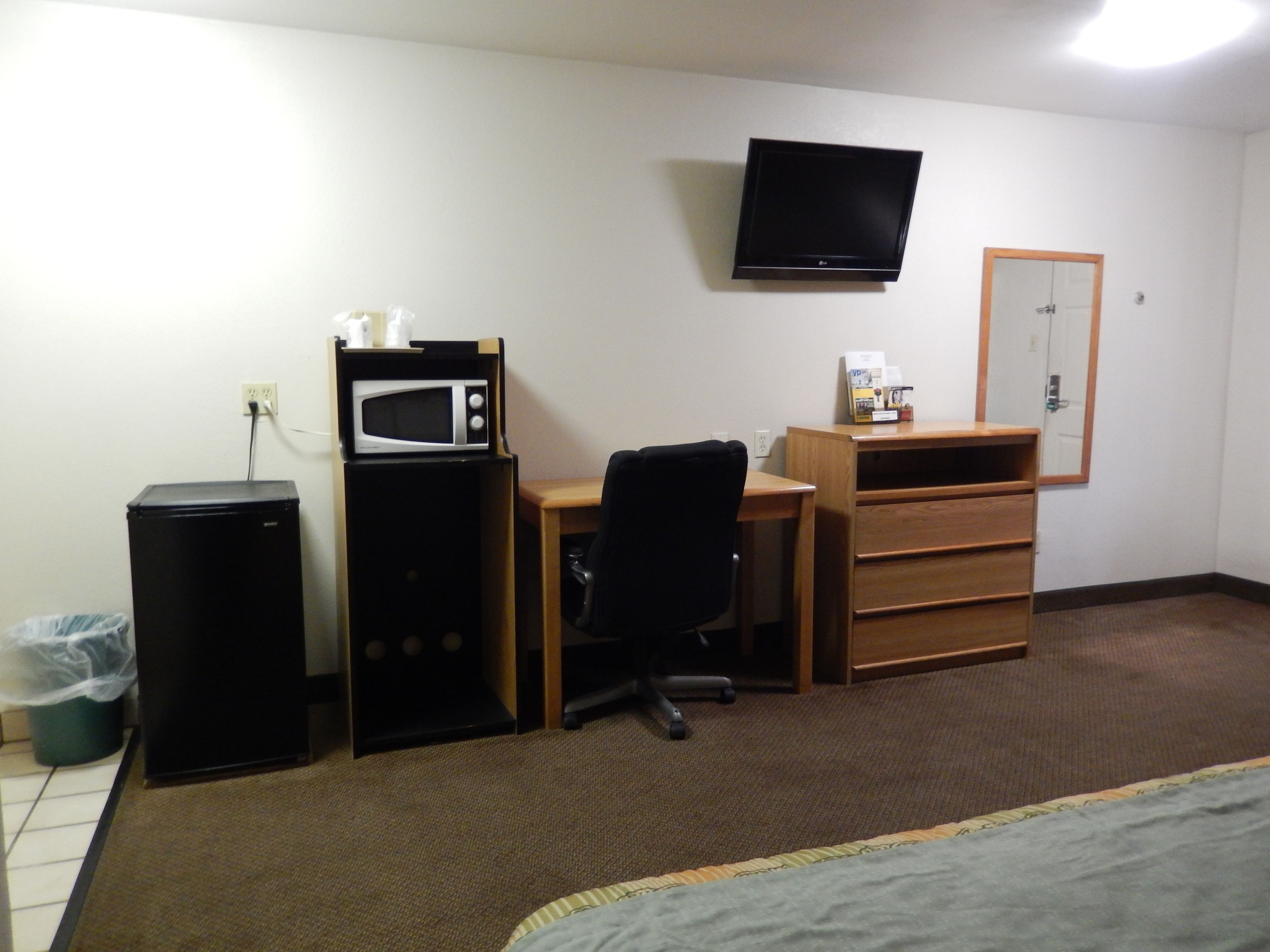 Una habitación de hotel pequeña con cama, escritorio, silla, microondas, refrigerador, televisor y cómoda de madera.