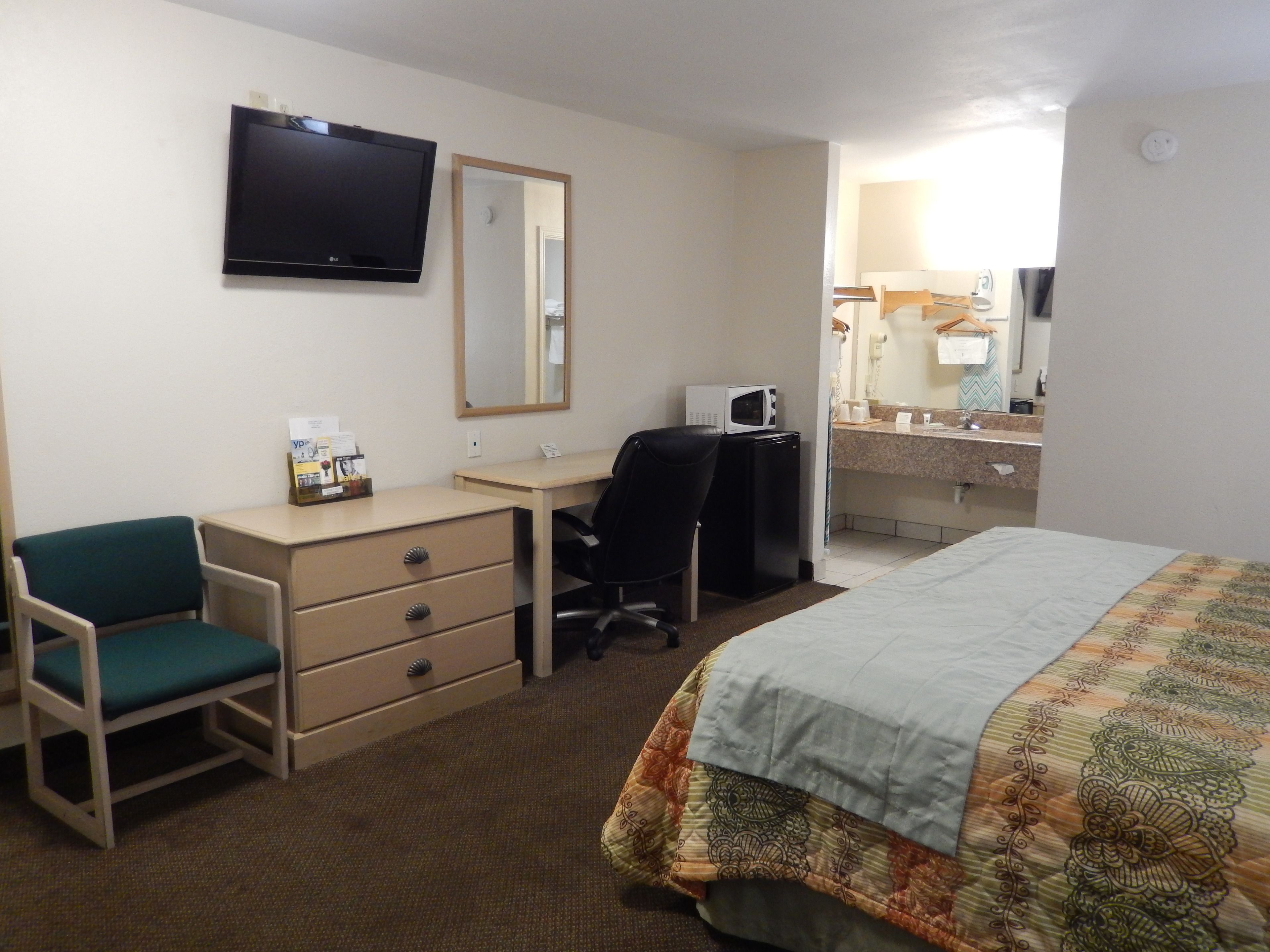 A hotel room with a bed, a desk with a chair, a television, a microwave, and a bathroom with a mirror and sink.