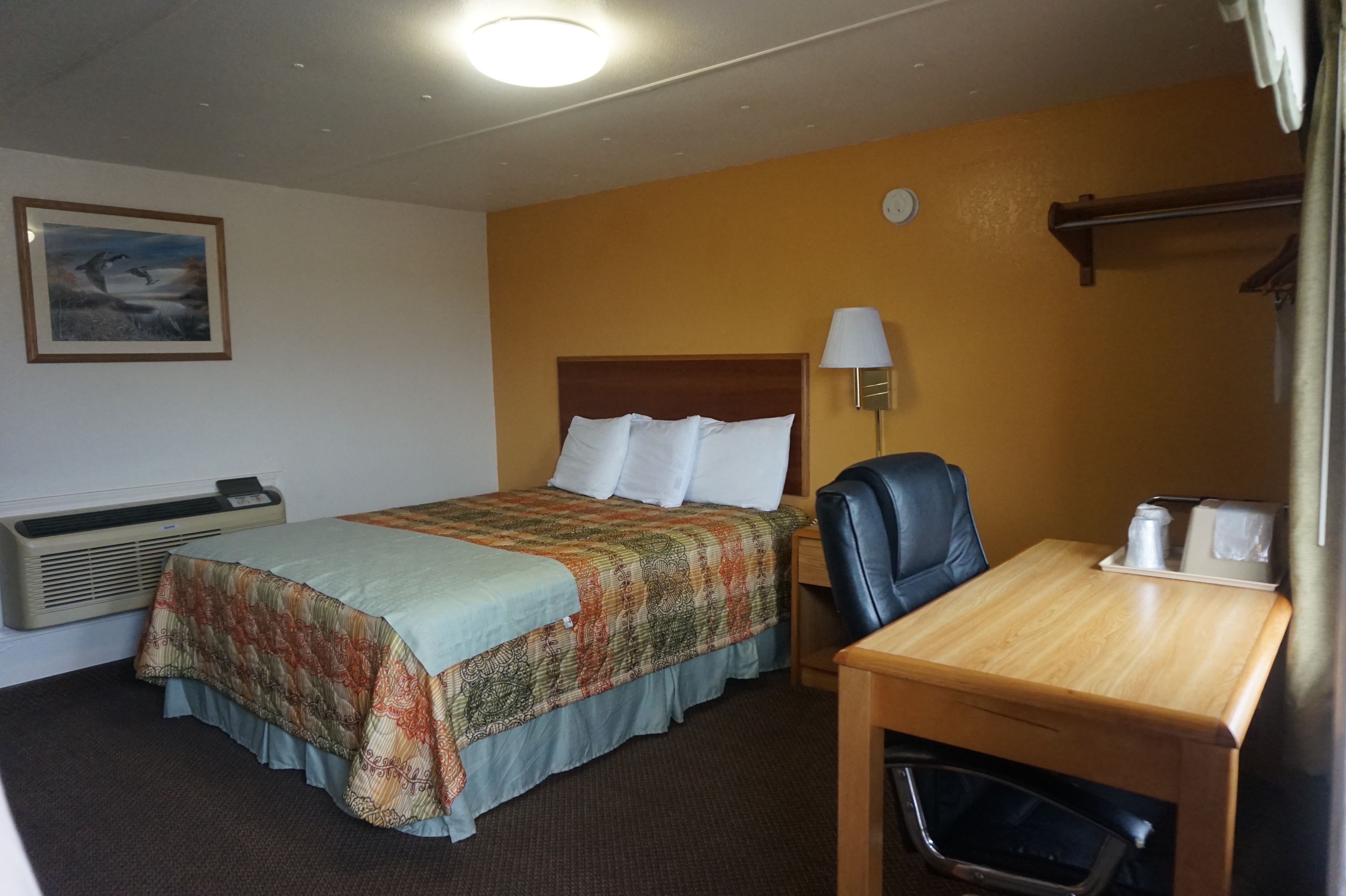Una habitación de hotel con cama, escritorio, silla, lámpara, reloj y un cuadro en la pared.