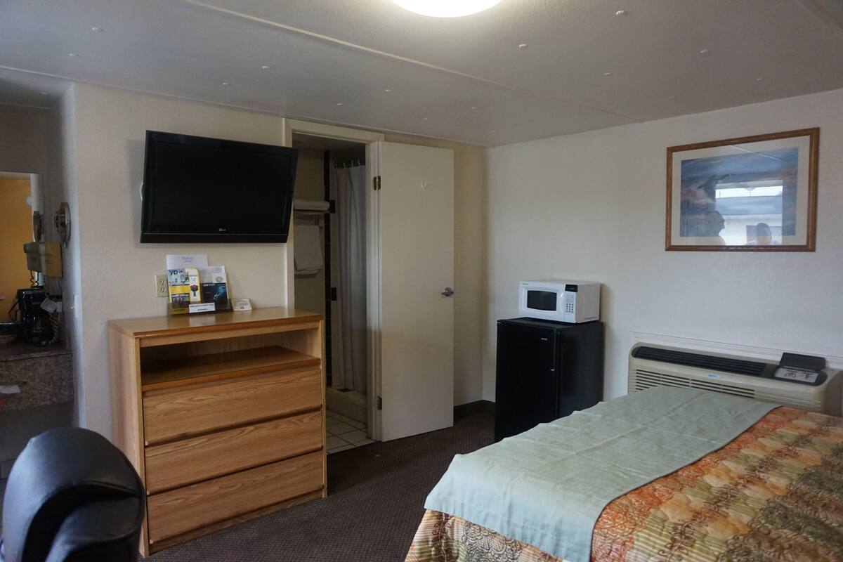 Une chambre d’hôtel comprenant un lit, un téléviseur fixé au mur, un four à micro-ondes, un radiateur, une commode en bois et une porte donnant sur une autre pièce.