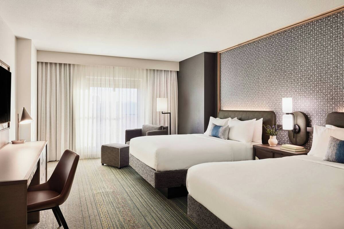 Ein Hotelzimmer mit zwei Betten, einem Schreibtisch, einem Stuhl, einem Fernseher und einem großen Fenster mit Vorhängen.